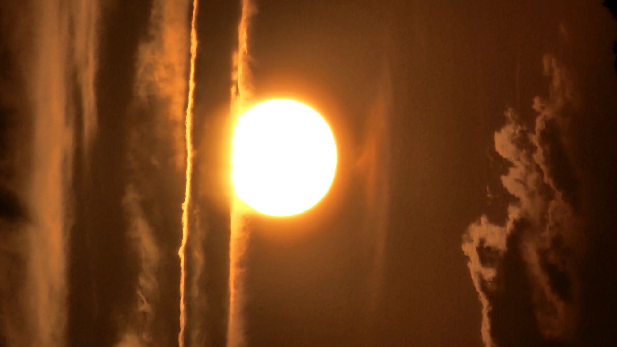 This media file depicts a close-up, vertical shot of the sun low in the sky, likely during sunrise or sunset. The sun appears as a bright, glowing white circle with a vibrant orange halo, dominating the center of the frame. The sky is a hazy orange, filled with dramatic clouds. On the left side, tall, wispy clouds with soft, blurred edges are illuminated by the sun. A sharp, vertical band of brighter light, possibly a ray of sunlight or a thin, intense cloud formation, runs down the left of the sun. On the right, darker, more textured clouds are visible, creating a contrast with the brighter elements. The overall mood is dramatic and awe-inspiring, with the intense light and cloud formations suggesting a significant atmospheric event. The location context provided is Asan-si, South Korea, which places the scene in an East Asian urban or semi-urban environment. There is no visible text in the image. The time of day is clearly indicated by the low angle of the sun and the quality of light. The weather appears to be partly cloudy with atmospheric haze.