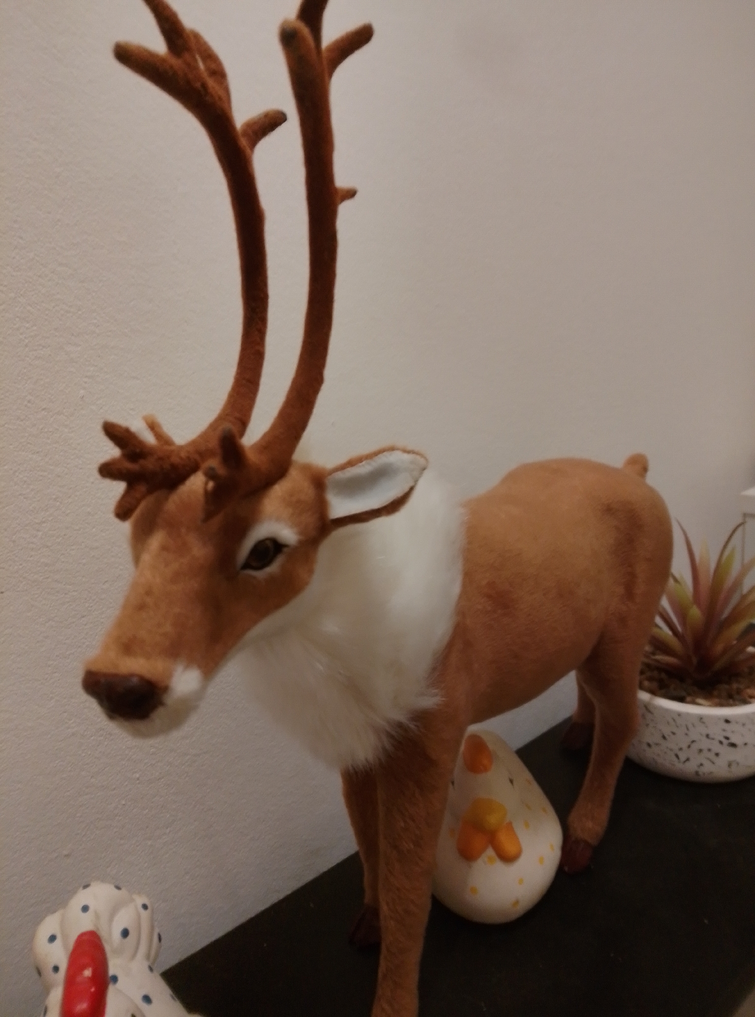 A stuffed toy reindeer with brown fur and large antlers stands on a dark surface against a white wall. A white fluffy ruff is around its neck. To the right of the reindeer, a potted plant with spiky, reddish-green leaves sits in a white speckled pot. In front of the reindeer's hind legs is a white, egg-shaped object with orange markings. To the lower left of the reindeer, a white ceramic figurine with blue polka dots and a red nose is visible. The overall scene appears to be an indoor decorative display. The location is not specified in the image.
