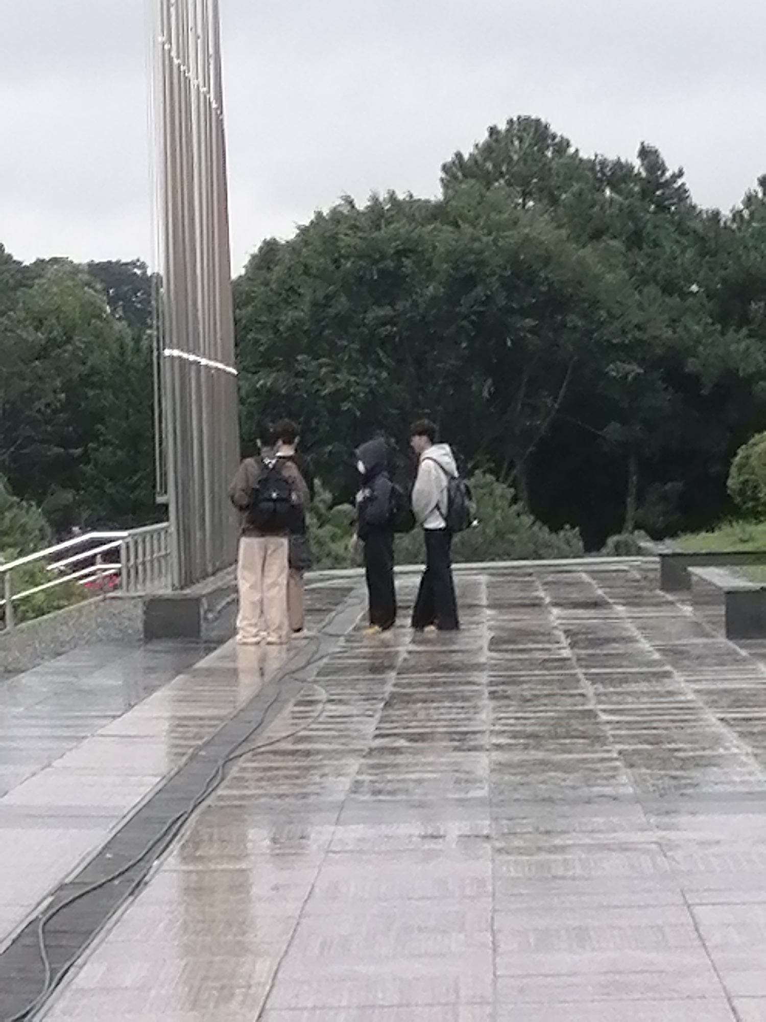 Four individuals are standing on a wet, paved plaza, with a large metal structure to the left. Three of the individuals are facing away from the camera, and one is partially facing to the side. The ground is reflective due to moisture, indicating it might be raining or recently rained. Trees form a dark background behind the plaza. The scene appears to be an outdoor public space. The metal structure resembles a monument or sculpture, possibly a flagpole due to its height and vertical lines. The people are casually dressed, with two wearing backpacks. The overall atmosphere is subdued, likely due to the overcast weather. This location is likely the Xuan Huong Lake area in Da Lat, Vietnam, given the presence of large open plazas and prominent outdoor monuments often found there.