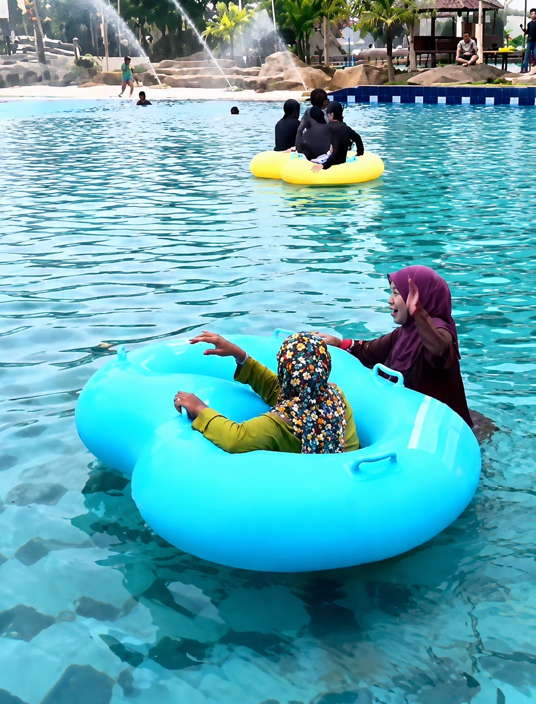 The image depicts a scene at a water park, likely in Eampat Kwalamencirim, Indonesia. In the foreground, two individuals are seated in a large, bright blue inflatable tube. One person, facing away from the camera, wears a patterned headscarf and a green long-sleeved shirt. Their hands are visible resting on the edge of the tube. To their right, another person wearing a purple headscarf and a dark top is partially visible, facing left and appearing to wave or reach out.
In the mid-ground, a group of at least three individuals, all wearing dark clothing, are seated in a yellow inflatable ring. They appear to be floating in the water.
The background features a water attraction with several water jets spraying water into the air, creating a misty effect. Several palm trees and other foliage are visible, along with rock formations. In the far background, a rustic wooden structure with a roof, possibly a seating area or shelter, is present. Other figures, including children and adults, are scattered throughout the water and on the paved areas surrounding the pool. The water is a clear, bright blue, with visible ripples and reflections. The overall atmosphere suggests a recreational setting on a sunny day.