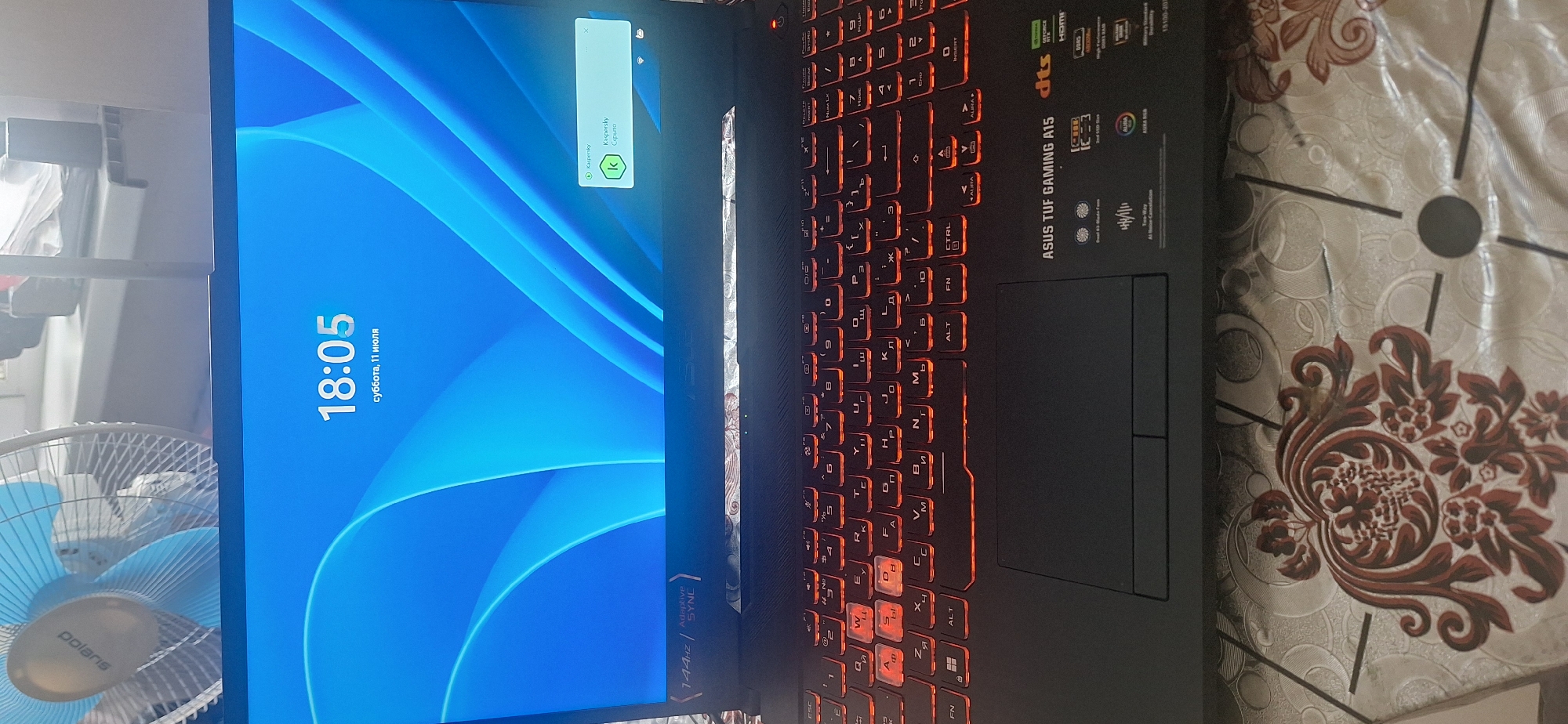 The image shows a laptop computer with a glowing red keyboard and a blue wallpaper displaying the time "18:05" and the date "суббота, 11 июля" (Saturday, July 11). A small window indicating "Kaspersky" is open in the upper right corner of the screen. The laptop appears to be placed on a patterned tablecloth. In the background, a blue fan is partially visible. The scene is not identifiable as being in Chunsky, Russia, or any other specific city.