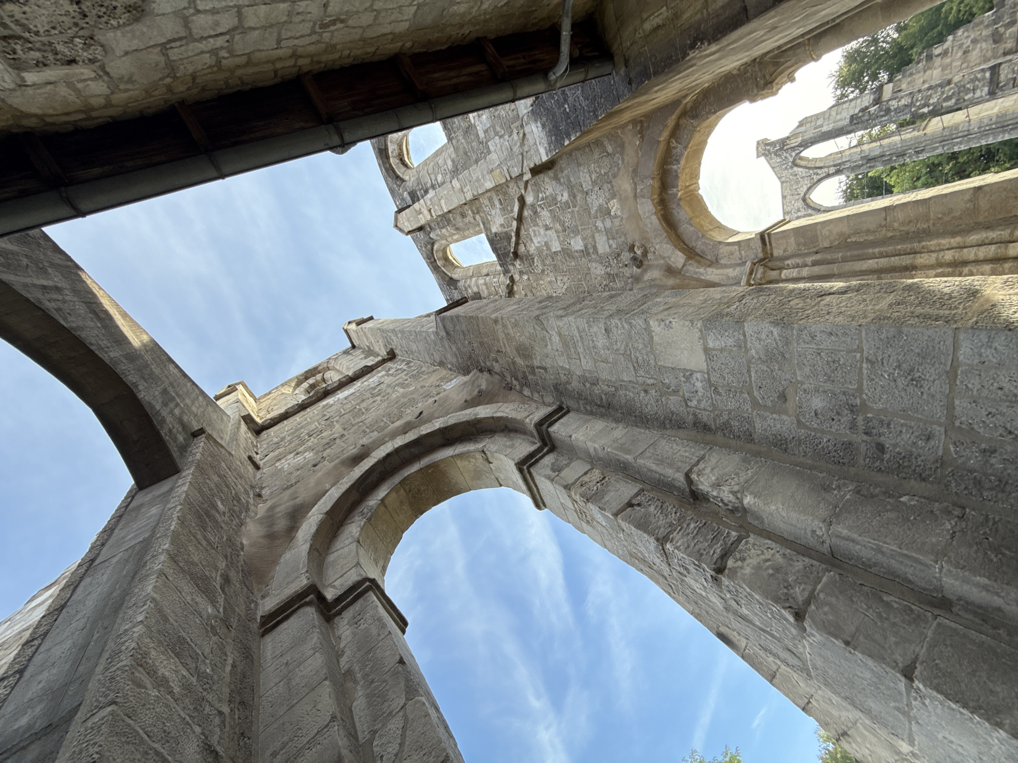 The image captures the imposing ruins of Walkenried Abbey in Walkenried, Germany. The perspective is low-angle, looking up at the weathered stone architecture against a bright blue sky with wispy clouds. Sunlight illuminates the scene, casting shadows and highlighting the textured surfaces of the ancient stonework. The foreground features a large, arched stone structure, appearing to be part of a wall or a gateway. Further back, the remnants of more arched openings and tall, slender columns of the abbey's former structure are visible, creating a sense of grandeur and decay. Lush green trees can be seen peeking through some of the openings in the background, suggesting the ruins are situated in a natural, possibly park-like setting. There are no people visible in the image. The overall impression is one of historical significance and the enduring beauty of a once-grand monastic complex.