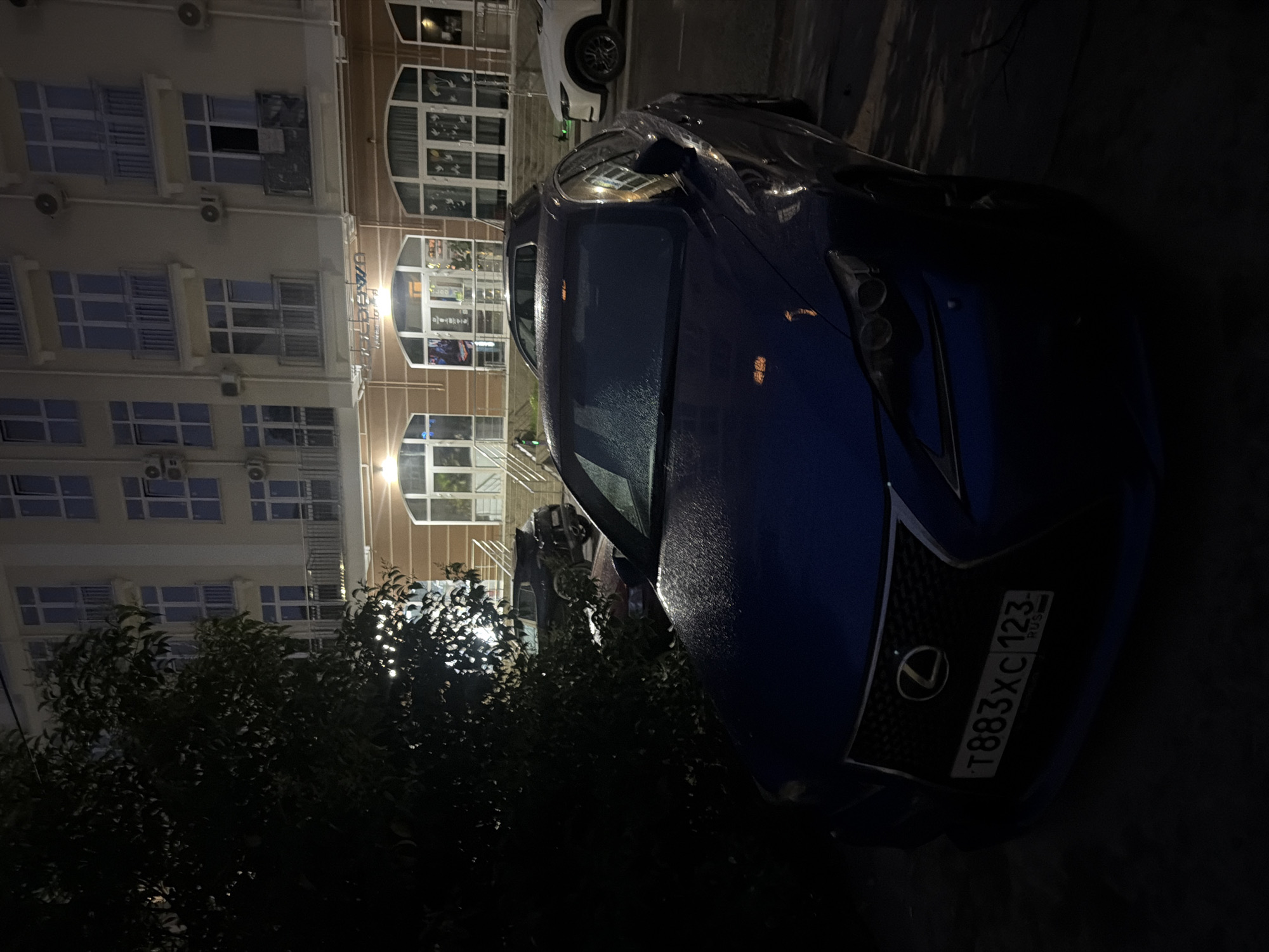 In Sochi, Russia, at night, a dark blue Lexus sedan is parked in front of a multi-story building with illuminated windows and balconies. The car's license plate reads "T883XC 123 RUS". A large, dark tree with visible leaves is to the left of the car. The ground appears wet, possibly from rain, with reflections visible on the car's hood and windshield. Another dark-colored vehicle is parked behind the Lexus, and a white SUV is visible further to the right. The building's facade is light-colored with dark window frames and multiple air conditioning units. The overall scene is dimly lit, with artificial light sources illuminating the building and parts of the vehicles.