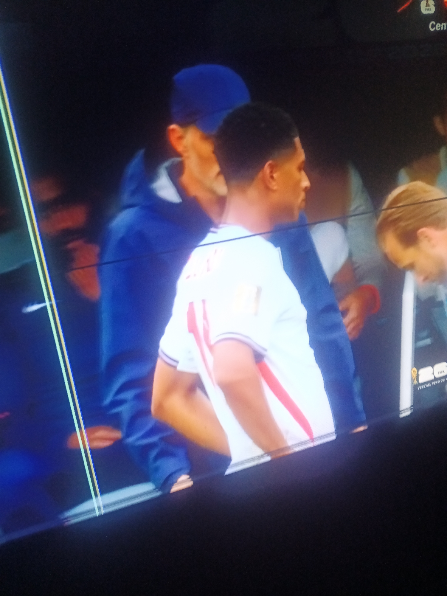 The media file shows a medium shot of several people in what appears to be a stadium or a large indoor venue. In the foreground, a young Black man wearing a white jersey with red stripes is visible from the side, his left arm bent and his head turned away from the camera. He has a light skin tone and a short haircut. Behind him, a man in a blue hooded jacket and a blue baseball cap is standing, his face partially obscured. Further back, other individuals are present, including a man with blond hair who is looking down. The setting seems to be indoors with dark background elements and some lighting. There are some vertical lines of light, possibly from stage lights or screen reflections, cutting across the image. In the lower right corner, there is a partially visible logo or graphic with text that is not clearly legible, but it appears to be related to a sports event, possibly featuring a trophy icon. The overall atmosphere is somewhat dim, suggesting it could be evening or an indoor event. No specific weather conditions are discernible.