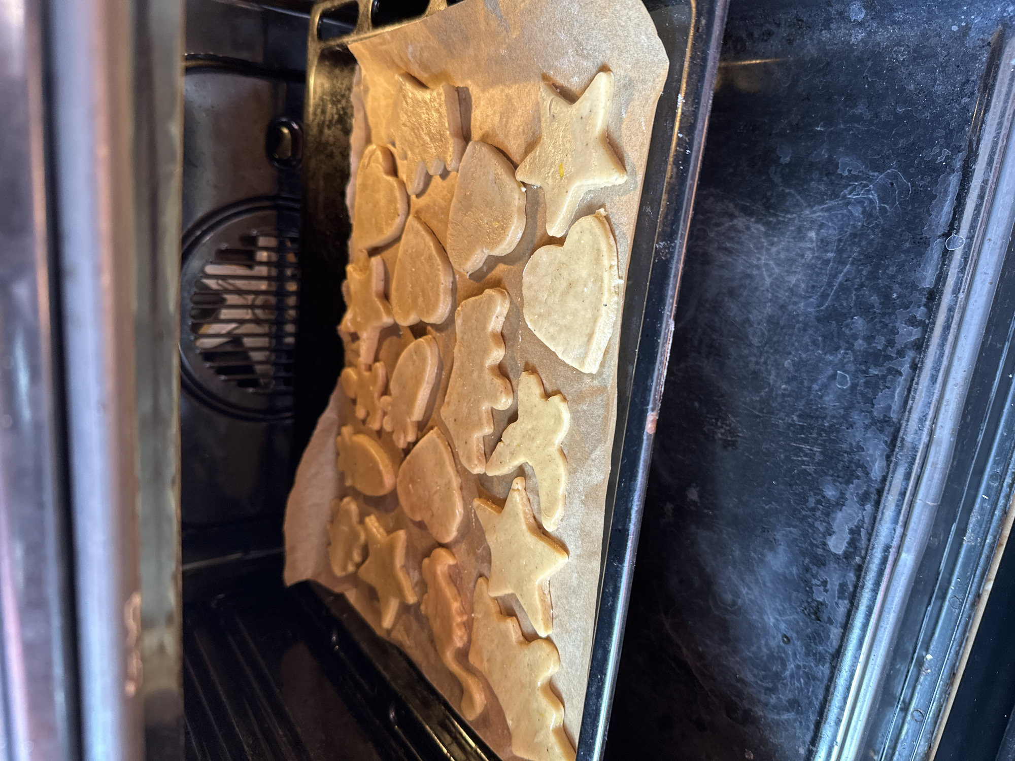 This is a close-up, interior shot of an oven with a baking sheet of unbaked cookies on the bottom rack. The cookies are cut into various shapes, including stars, hearts, and animal figures. They are arranged on parchment paper, which covers the baking sheet. The oven's heating element and fan are visible in the background. The scene is indoors, likely in a kitchen. There are no people or animals present in the image. The location is not specified as Moscow, Russia, as there are no visual cues to support this claim.