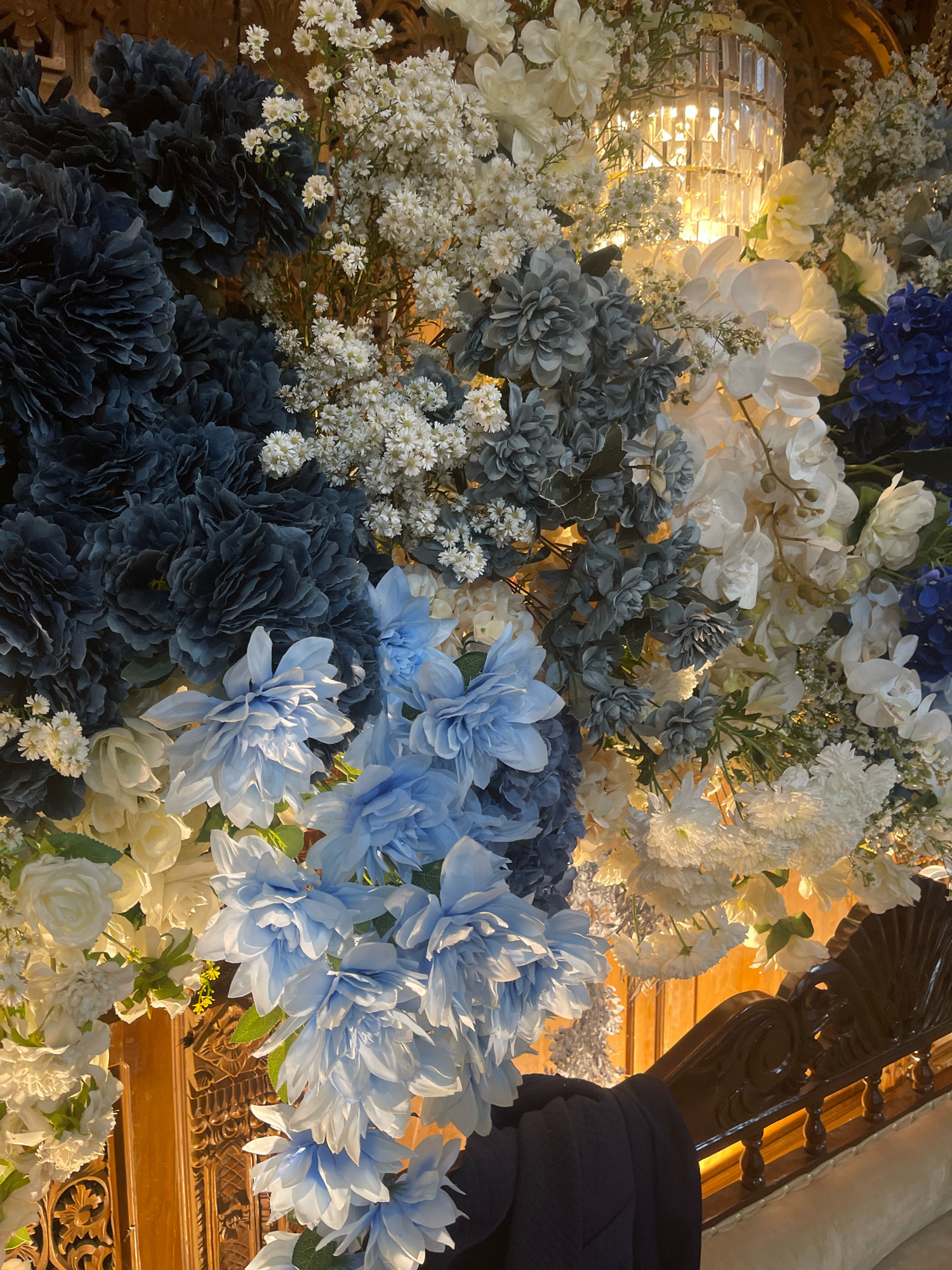 The image displays a densely packed floral arrangement featuring a variety of artificial flowers in shades of dark blue, gray, light blue, white, and cream. The flowers are arranged in a wall-like display, with large, textured blooms dominating the foreground and midground, interspersed with smaller, delicate white flowers. In the upper central portion of the image, a portion of a crystal chandelier with a warm, yellow light source is visible, suggesting an indoor event or decorated space. The floral arrangement is positioned in front of a wooden structure that appears to be part of an ornate staircase or balcony railing. The wood is dark and intricately carved. The overall impression is one of opulent decoration, likely for a wedding or formal event. The image context provided indicates that this scene is located in the city of Bagusan, Indonesia, though the image itself does not contain any specific geographical markers to confirm this. No people are visible in the image.