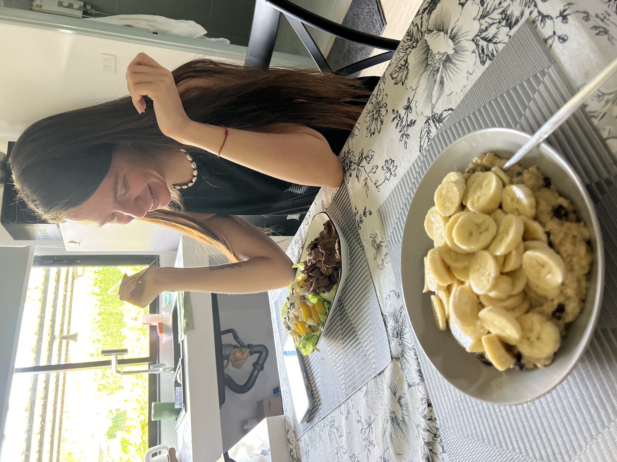 The image depicts a woman sitting at a table indoors. She has long brown hair and is wearing a dark-colored top. She is smiling and appears to be interacting with food on the table. A bowl of oatmeal topped with sliced bananas is prominently positioned. Next to it, there is a plate containing a salad with meat, mangoes, and greens. The table is covered with a floral-patterned tablecloth and a striped placemat. A kitchen sink area with a window providing an exterior view with trees and greenery is in the background. The image was taken in บ้านบางรักษ์, Thailand.