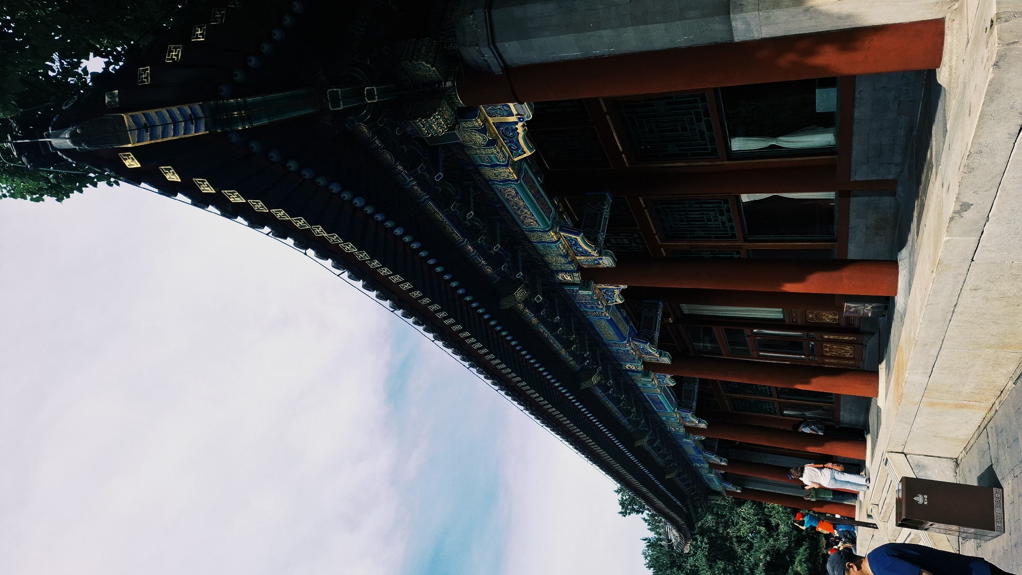 The image shows a low-angle, partially obscured view of a traditional Chinese building in Haidian District, China. The building features red pillars, intricately painted eaves in blue, green, and gold, and latticed windows. Several people are visible in the bottom right, including at least one person wearing a white shirt and jeans. A trash receptacle stands nearby. The foreground to the left is mostly sky with light clouds. Trees are visible at the edges of the building.