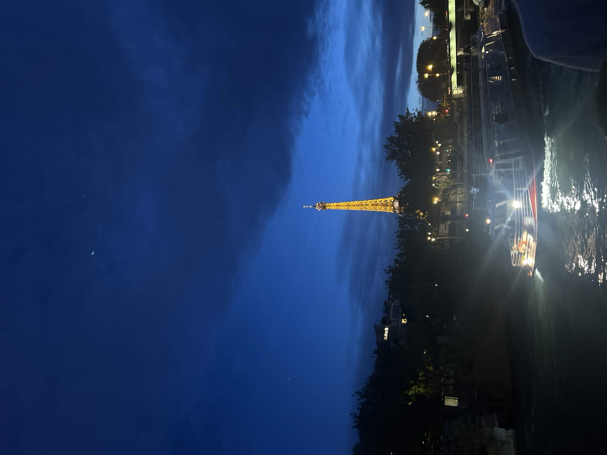 The image depicts the Eiffel Tower illuminated against a blue evening sky in Paris, France. The tower is positioned slightly off-center in the frame. To the right of the tower, trees and buildings line the Seine River. Light reflects on the water's surface, and a boat with illuminated windows is visible. The sky transitions from a darker blue on the left to lighter hues near the horizon.
