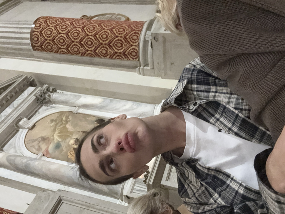 The image captures the interior of the Chiesa di San Zaccaria in Venice, Italy. In the foreground, a young adult female is centered, looking upwards and to the right with a pouting, "duck face" expression. She has dark hair pulled back and wears a white t-shirt under an open dark blue and white plaid button-up shirt. Partially visible to her right is the head and shoulder of an individual with blonde hair, wearing a brown ribbed sweater. A portion of another person's light-colored hair is visible to her left. The background features classical Baroque architecture, including tall, veined grey and white marble columns. Between two columns, an arched niche contains a fresco or painting depicting celestial figures amidst clouds, rendered in muted tones. Above the niche, white architectural molding is visible. To the far right, a cylindrical structure, possibly a pillar or a section of a pulpit, is covered in a rich red brocade fabric adorned with repetitive gold stylized heart or leaf patterns. The lighting within the space appears dim, emphasizing the architectural and artistic details.