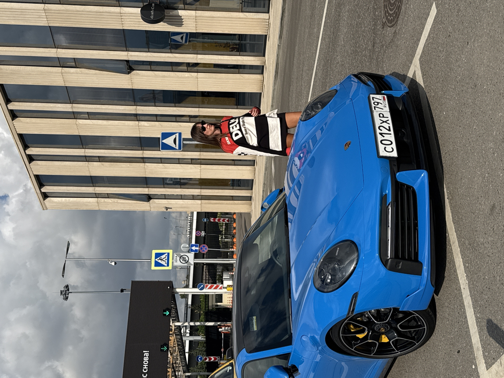 A woman stands next to a blue Porsche 911 in Moscow, Russia. The building behind them features a modern architectural style with a facade of vertical, beige-colored panels and dark windows. The woman is wearing a black mini-skirt and a long-sleeved shirt with red, white, and black stripes, featuring the "DEUX" logo and text that reads "House of Simple Pleasures." She is also wearing sunglasses. The car has a prominent license plate that reads "C012XP 797 RUS". In the background, there are several traffic signs, including a pedestrian crossing sign and a speed limit sign, as well as lampposts and what appears to be an electronic billboard with Russian text. The scene is set on an asphalt road, suggesting an urban environment, possibly near an entrance or exit point, given the presence of traffic control elements. The sky is overcast with dramatic clouds.