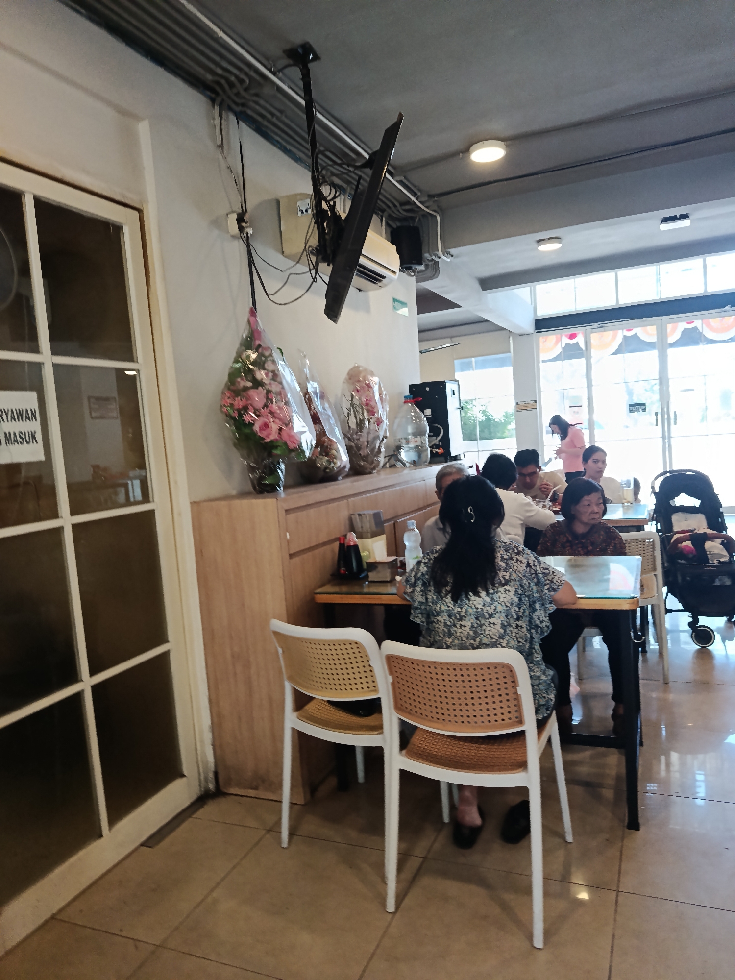 The image depicts the interior of a restaurant in Surabaya, Indonesia. The scene is brightly lit and appears to be midday. In the foreground, there are several tables with chairs, some occupied by diners. One woman in a floral-patterned shirt is seated at a table, facing away from the camera. Another woman with dark hair and a patterned top is seated at the same table. Further back, more people are seated at tables, engaged in conversation and dining. A baby stroller is visible to the right. The restaurant has large windows with decorative elements, suggesting a public dining establishment. The decor includes floral arrangements and what appears to be a television mounted on the ceiling. The overall impression is of a casual and moderately busy restaurant environment.
