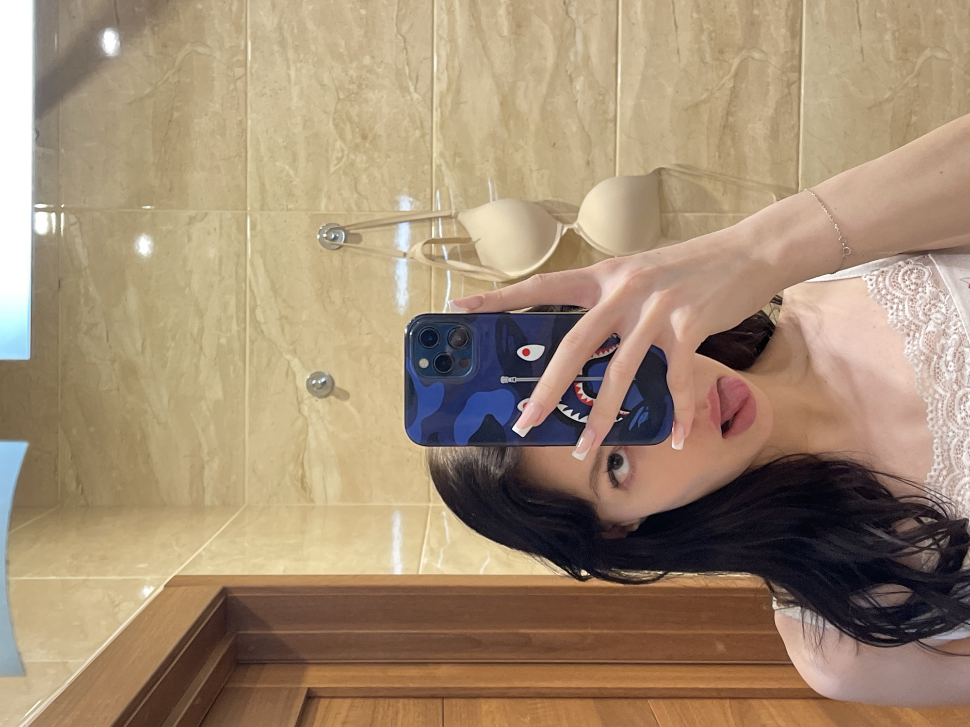 A young woman with long, dark hair takes a selfie in a bathroom with marble-patterned walls. She is wearing a white lace-trimmed top and a thin silver bracelet. She holds a blue phone with a shark graphic in her right hand, obscuring part of her face. Her left eye is visible, and her tongue is sticking out. Two beige bras hang on hooks on the wall behind her. The room appears to be a private residence in Khimki, Russia.