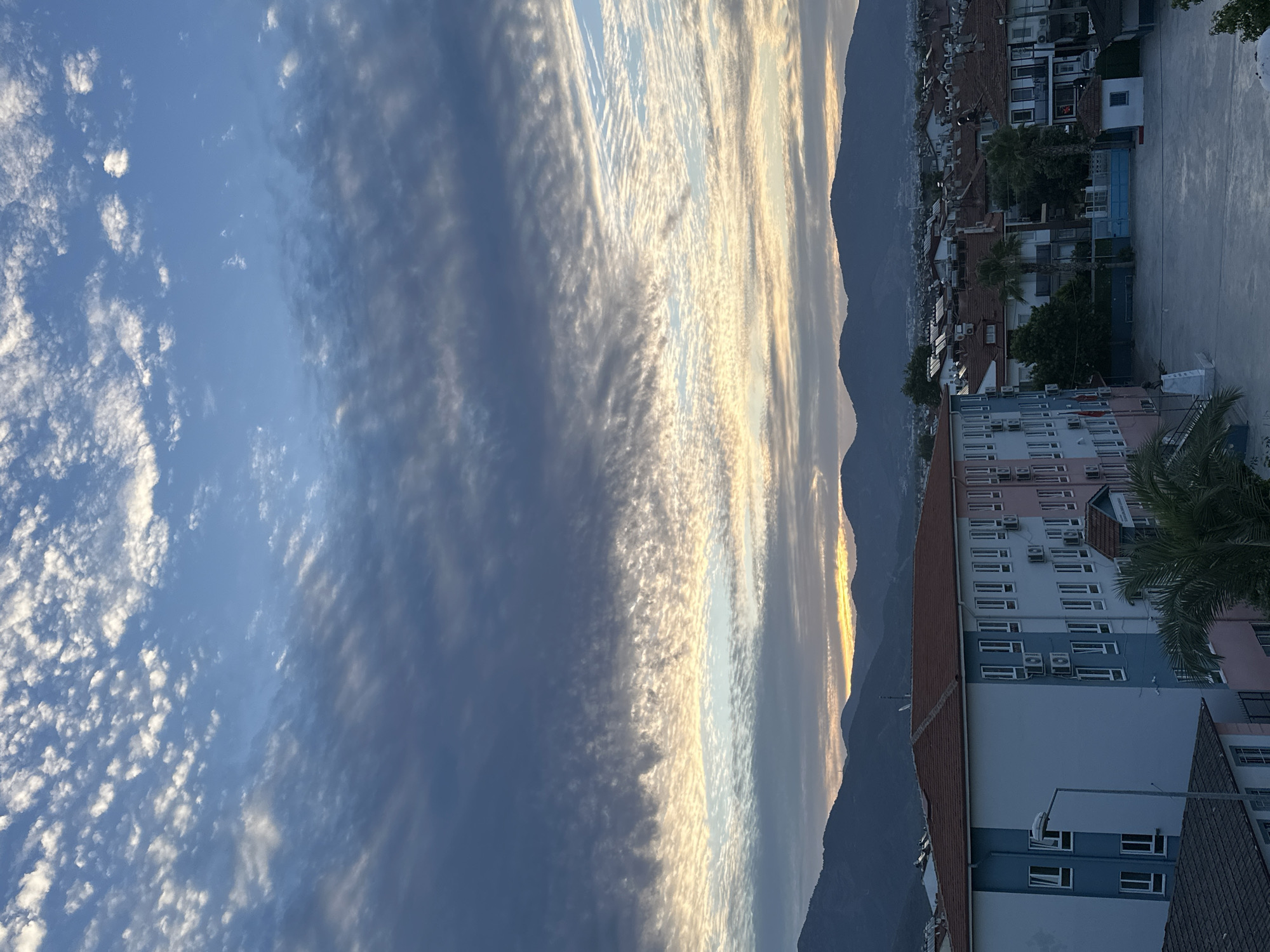 The image captures an exterior view of a school building in Fethiye, Turkey, under a dramatic, cloud-filled sky. The school building itself is a multi-story structure with a reddish-brown tiled roof. The facade is painted in segments of light blue, white, and pink, with rows of windows visible on each level. Air conditioning units are mounted on the exterior walls. A lamppost stands to the left of the building, and a palm tree is visible in the foreground to the right of the main building entrance.
The schoolyard in front of the building is paved and empty, suggesting it is either early morning or late afternoon. In the middle ground, residential buildings with similar tiled roofs are visible, forming a typical urban landscape. Beyond the town, a range of dark, silhouetted mountains forms the backdrop.
The sky is dominated by a vast expanse of clouds, displaying a variety of textures and colors. Streaky, wispy clouds intermingle with denser formations, creating patterns of light and shadow. The setting sun or rising sun is not directly visible, but the lower part of the sky shows hues of yellow and orange, indicating the presence of sunlight breaking through or reflecting off the clouds. The overall impression is one of a serene, late-day scene in a coastal Turkish town.