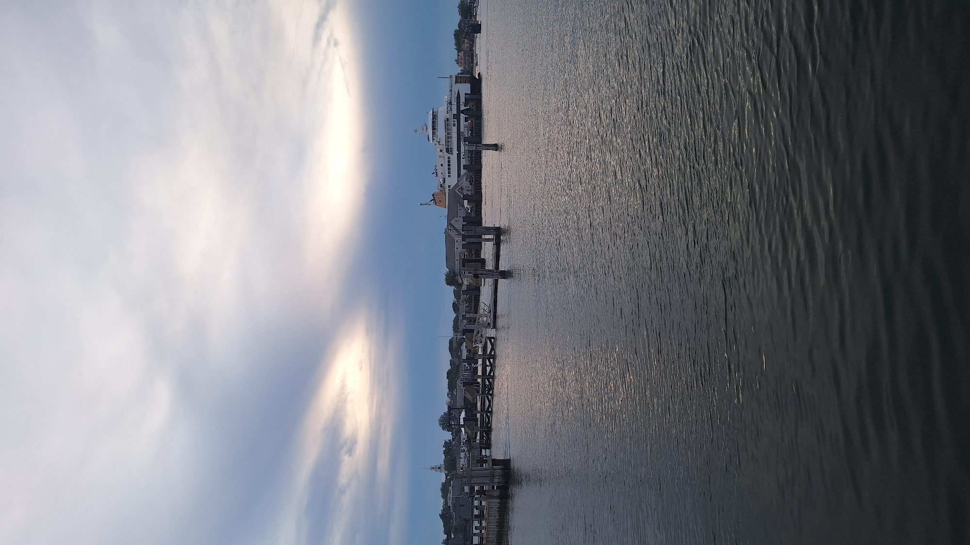 The image depicts a waterfront scene in Nantucket, Massachusetts, USA. A large ferry is docked at the terminal, with several buildings and a white steeple visible in the background, suggesting a town or village setting. Several wooden piers and pilings extend into the water in the foreground. The sky is overcast with streaks of light breaking through the clouds, indicating either dawn or dusk. The water is calm, with ripples reflecting the muted light. There are no people visible in the image. The overall atmosphere is serene and quiet, characteristic of a coastal town.