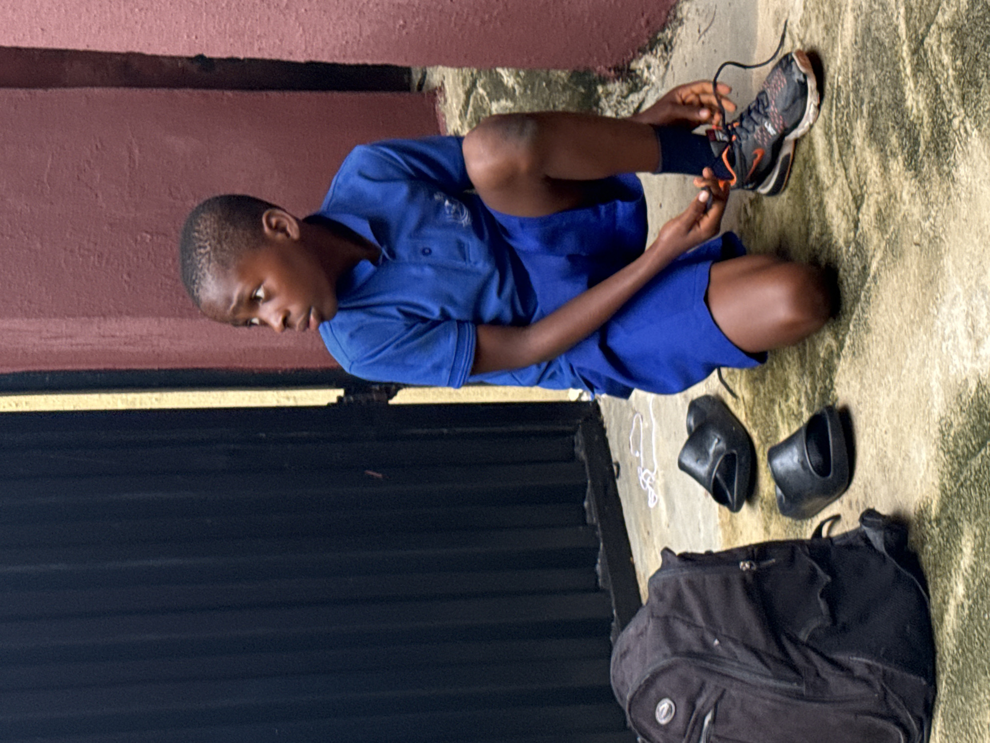A young person in a blue collared shirt and matching shorts kneels on a weathered concrete surface tying the laces of a dark athletic shoe with orange accents. Beside them are a pair of black sandals, a black backpack, and stray white laces. The scene is framed by a reddish-brown wall on the left and a black metal gate in the foreground. The location is likely a schoolyard or similar public space within the city of Ado, Nigeria.