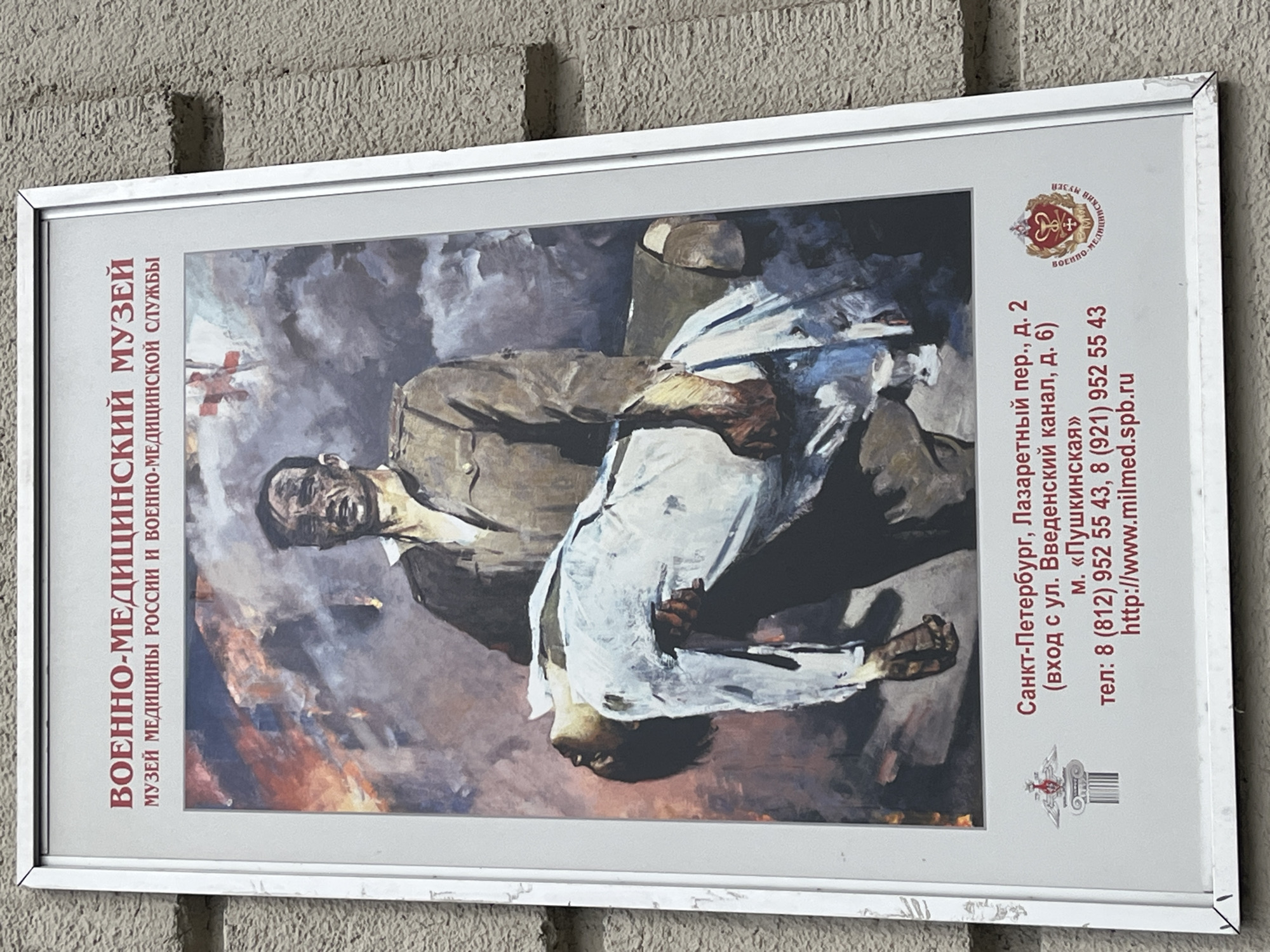 An outdoor poster, framed in silver metal, is displayed against a textured, light-colored wall, likely concrete or stucco. The poster features a painting depicting a dramatic scene. In the painting, an adult male, possibly a soldier or medic, dressed in a brown uniform jacket, is shown carrying a limp individual in light-colored (white/light blue) clothing. The carrier looks upwards with a strained or distressed expression, suggesting effort and emotion. The person being carried appears to be a casualty, possibly injured or deceased. The background of the painting is rendered with broad, sweeping brushstrokes in muted, chaotic colors, hinting at a scene of conflict, fire, or distress.
To the left of the painting, running vertically, is the museum's name in Russian: "ВОЕННО-МЕДИЦИНСКИЙ МУЗЕЙ" (Military Medical Museum) and "МУЗЕЙ МЕДИЦИНЫ РОССИИ И ВОЕННО-МЕДИЦИНСКОЙ СЛУЖБЫ" (Museum of Medicine of Russia and the Military Medical Service). To the right, also running vertically, are contact and location details: "Санкт-Петербург, Лазаретный пер., д. 2" (St. Petersburg, Lazaretny Lane, building 2), with an alternate entrance specified as "(вход с ул. Введенский канал, д. 6)" (entrance from Vvedensky Canal street, building 6). It also mentions the metro station: "м. «Пушкинская»" (Metro station "Pushkinskaya"). Two phone numbers are listed: "тел: 8 (812) 952 55 43, 8 (921) 952 55 43," and the website "http://www.milmed.spb.ru." Two logos are visible: a circular emblem with a caduceus-like symbol at the top right and a smaller, more stylized emblem at the bottom right. The poster itself shows minor wear, particularly on the frame.