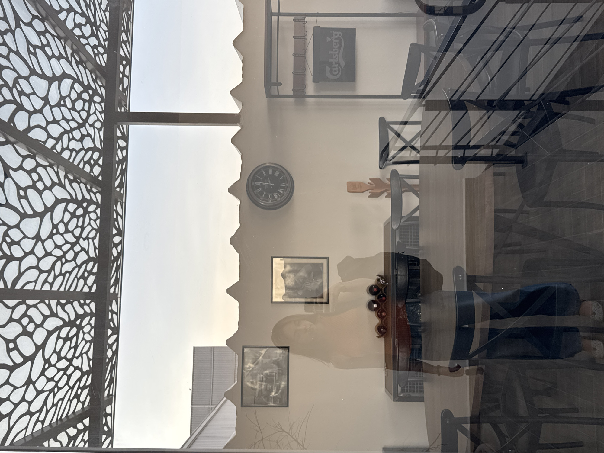 The image, likely taken indoors in San Bernardino Tlaxcalancingo, Mexico, shows an interplay of reflections and direct views. On the left, a lattice structure is visible, suggesting a window or partition. Outside, the sky is visible with a building's corner towards the bottom. The main interior reflects off the glass. Inside, we see a wall with decor including a Carlsberg sign, a clock, and framed pictures. There is a person reflecting in the glass, along with furniture like chairs and a wine rack.