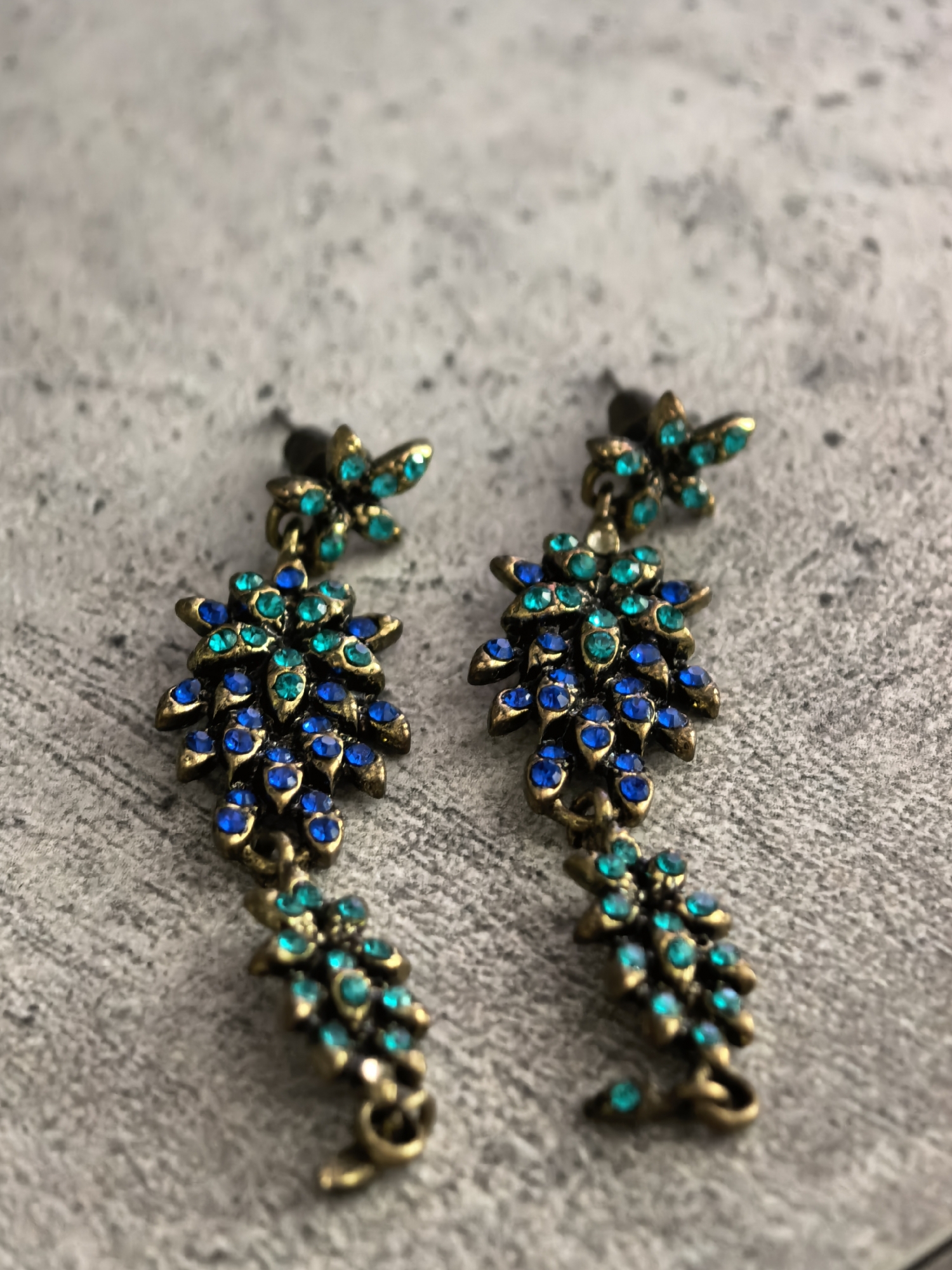 A pair of antique-style dangle earrings, featuring a gold-toned metal setting adorned with faceted blue and teal rhinestones, are placed on a textured gray surface. The earrings are designed with a floral or leaf motif, with the stones clustered to resemble petals or leaves. The surface appears to be concrete or stone, with subtle variations in color and texture. The lighting is soft, casting gentle shadows. There are no people or any indications of a specific location within Sterlitamak, Russia.