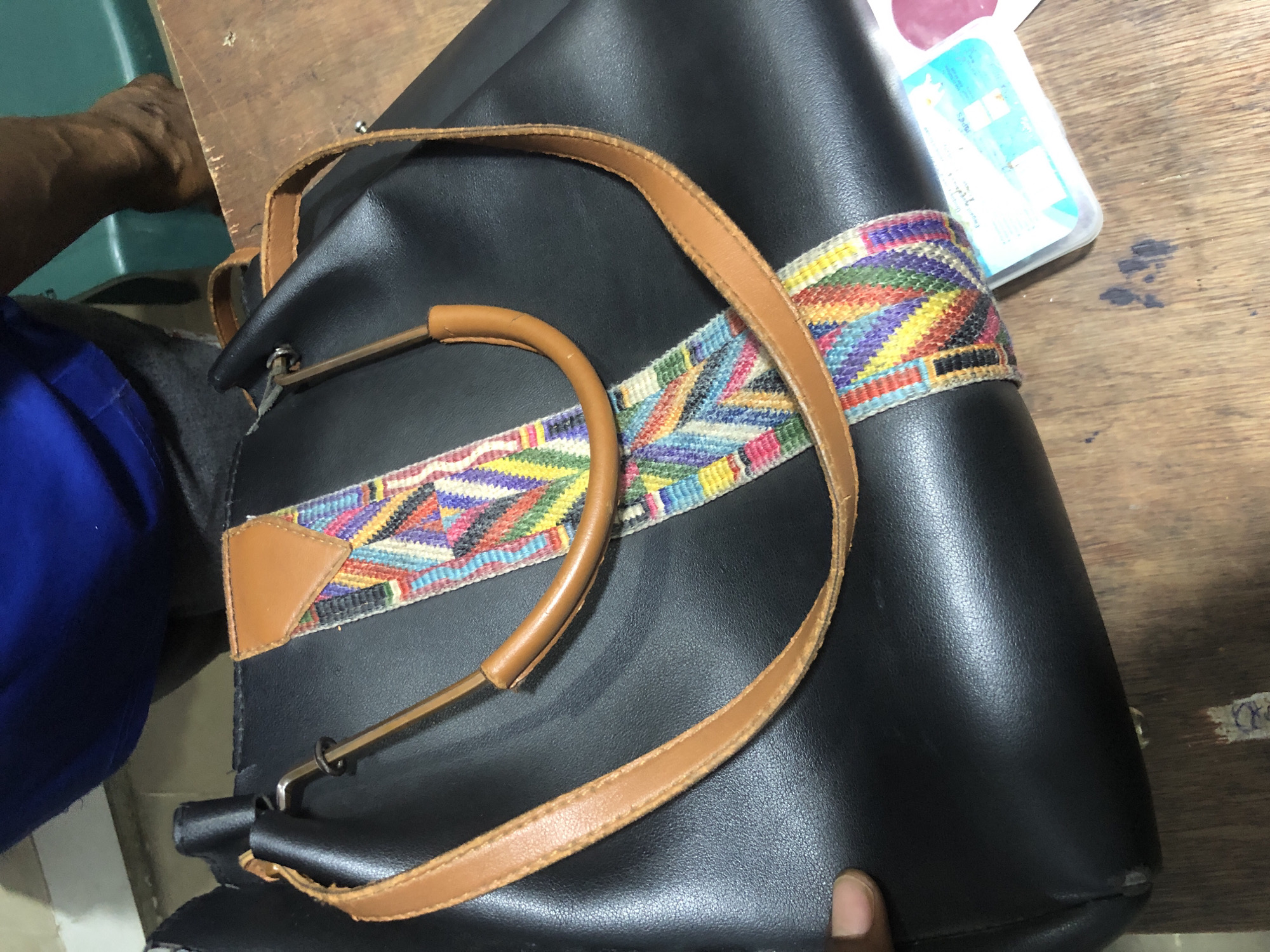 A black leather tote bag with tan leather straps and a colorful woven strap across the front is displayed on a wooden table. The bag is partially obscured by a blue shirt worn by an unseen individual, whose right arm and hand are visible near the back of the bag, resting on a green surface. A small plastic container with white contents and a pink object are placed near the lower right corner of the bag. The scene is lit from above, casting subtle shadows. The table's surface is worn and appears to be made of aged wood. The background is a blur of muted colors, suggesting an indoor environment. The image quality is decent, with clear focus on the bag and its details. The overall impression is that of a casual, everyday setting, possibly in a home or a shop in Ado, Nigeria, given the context.