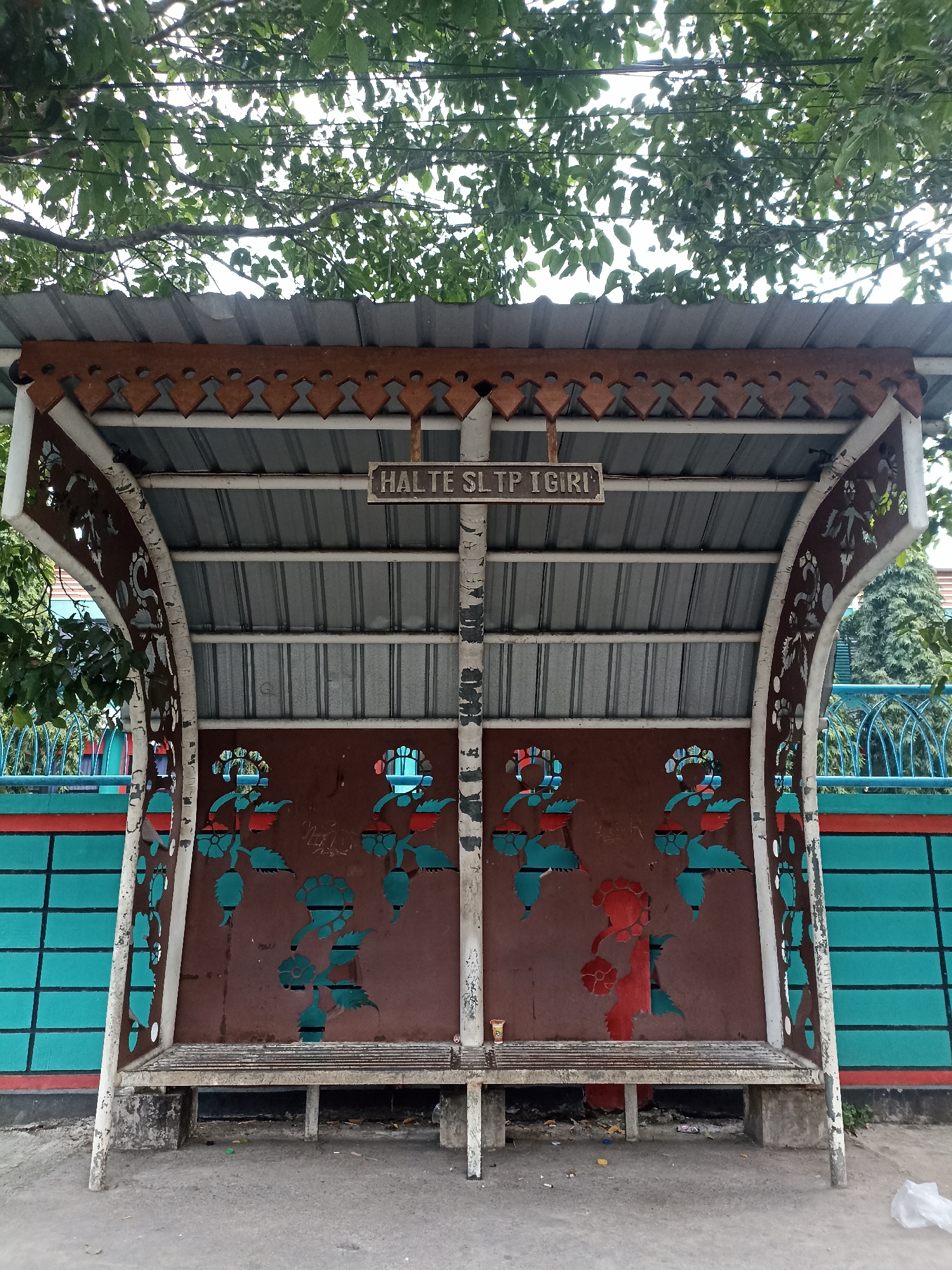 This is a medium shot, eye-level photograph of a bus stop shelter located in Mojopanggung, Indonesia. The shelter has a corrugated metal roof with a decorative, sawtooth-edged wooden trim along the front. Two white support poles with ornate, carved patterns are visible. The back wall of the shelter is painted a reddish-brown and features cut-out decorative patterns of flowers and leaves in teal and red. Above the back wall, a sign reads "HALTE SLTP IGIRI." Two weathered, slatted benches are situated inside the shelter. In front of the shelter is a concrete ground, littered with small pieces of trash, including what appears to be cigarette butts and a plastic wrapper. A teal-colored wall with a horizontal red stripe runs along the left side of the frame, partially obscuring a blue, decorative metal fence in the background. Lush green trees with dense foliage are visible above and to the left of the shelter, with power lines running through the branches. The overall impression is one of a somewhat neglected but functional public space.