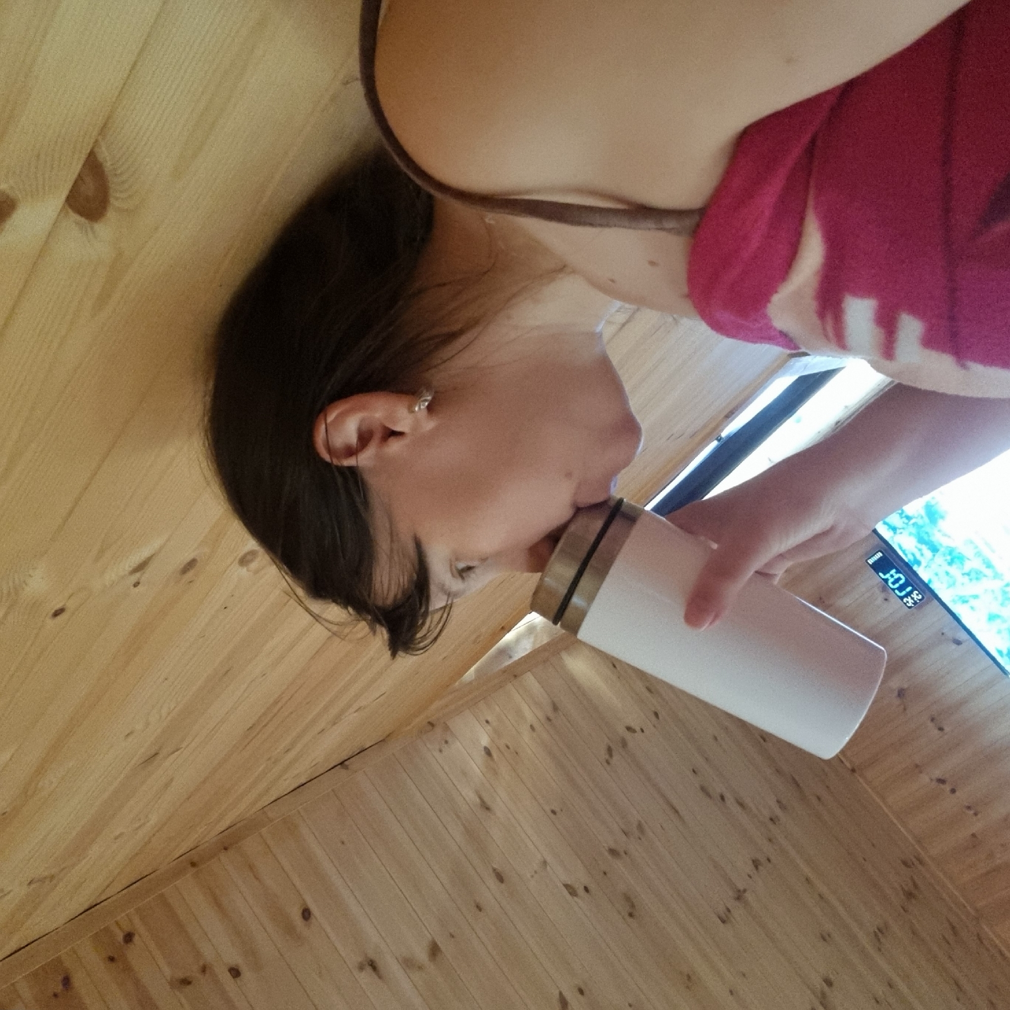 In Dunnae-myeon, South Korea, a woman is seen from the chest up, holding a white travel mug to her lips. She is looking to the left, with her dark hair cascading over her shoulder. She is wearing a pink and white patterned top. The background consists of wooden walls that appear to be part of a slanted ceiling, suggesting an interior space. A portion of a television screen is visible at the bottom of the image. The lighting is natural, casting soft shadows.