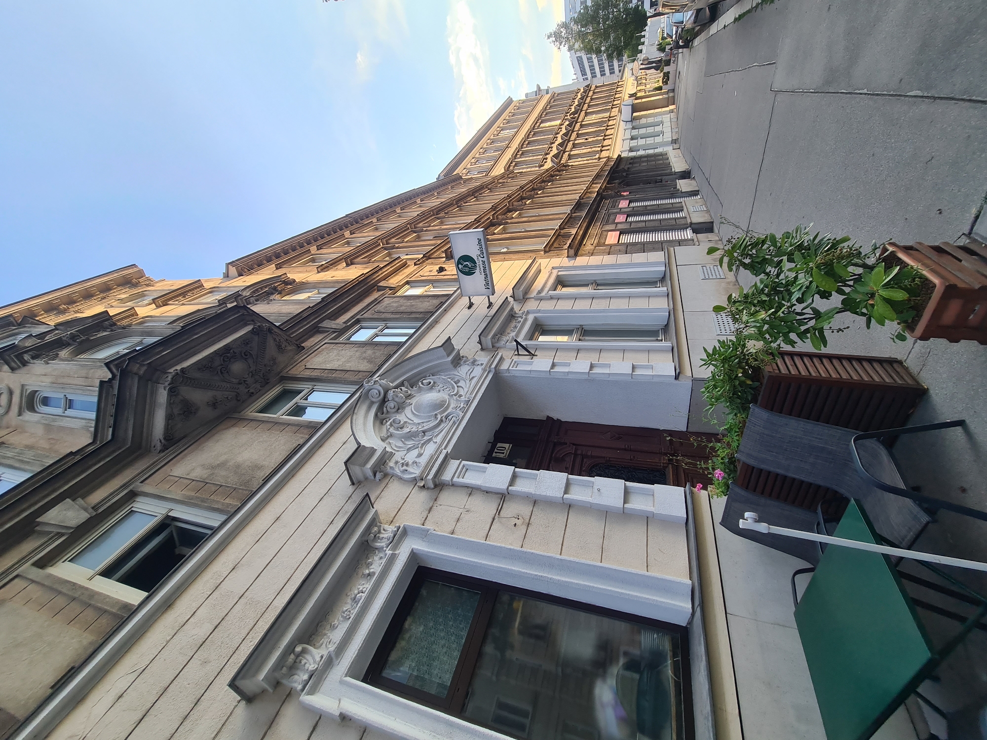 The image captures a street view in Vienna, Austria, with a focus on the facade of a building that houses a Vietnamese restaurant. The building's architecture features ornate detailing around the entrance and windows, with a neutral-toned facade. A sign for "HOANG LONG VIETNAMESE CUISINE" is prominently displayed on the building's upper level. In the foreground, an outdoor seating area is set up with a green table and two dark chairs, adjacent to a wooden planter filled with green foliage. The sidewalk runs alongside the building, leading towards other establishments and a street with parked cars. The sky is clear and blue, suggesting it is daytime. The scene depicts a quiet urban setting with a commercial establishment and outdoor dining potential.