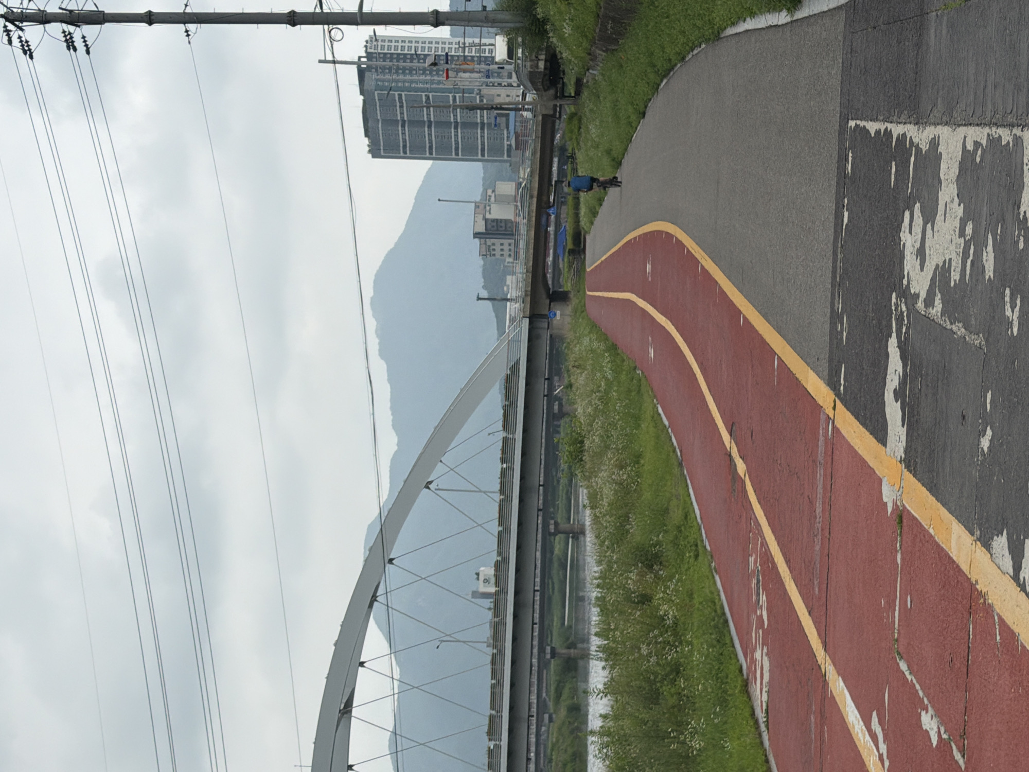 The image depicts a paved bike path with a red lane and a grey lane, separated by yellow lines. The path curves to the right in the foreground. A lone cyclist wearing a blue shirt and a dark helmet is riding away from the camera on the grey lane. To the left of the path is a river with grassy banks dotted with white wildflowers. Beyond the river, a multi-lane bridge crosses the water, and further in the distance, a larger, modern arch bridge spans the river. In the background, several buildings, including a tall, modern high-rise, are visible against a backdrop of misty mountains. Overhead, power lines crisscross the cloudy sky. The scene is consistent with the bike paths often found along rivers in urban areas of Dongducheon-si, South Korea, offering recreational opportunities amidst an urban and natural landscape.