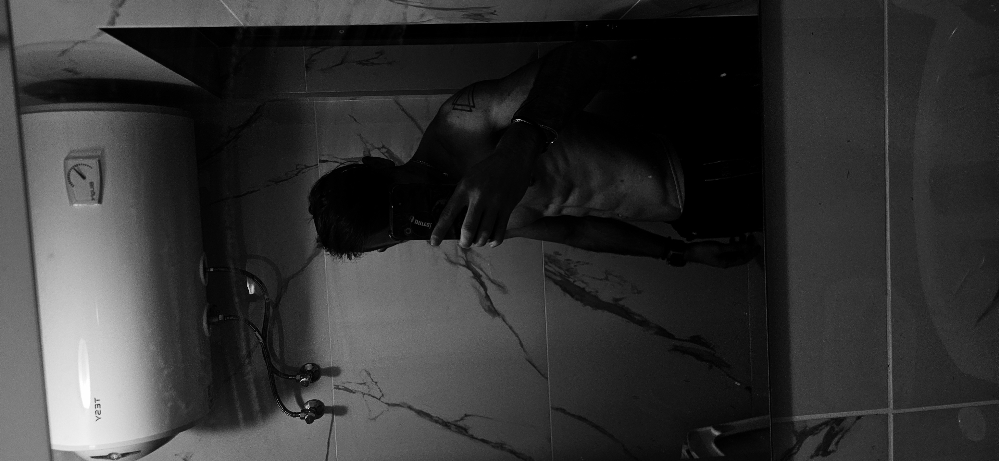 The image depicts a bathroom setting, likely in Plovdiv, Bulgaria, showing a man taking a picture of himself in the mirror. The scene includes a water heater fixed on the wall, with visible plumbing. The man, shirtless, is holding his phone to capture the reflection. The bathroom is tiled, featuring a marble-like pattern.