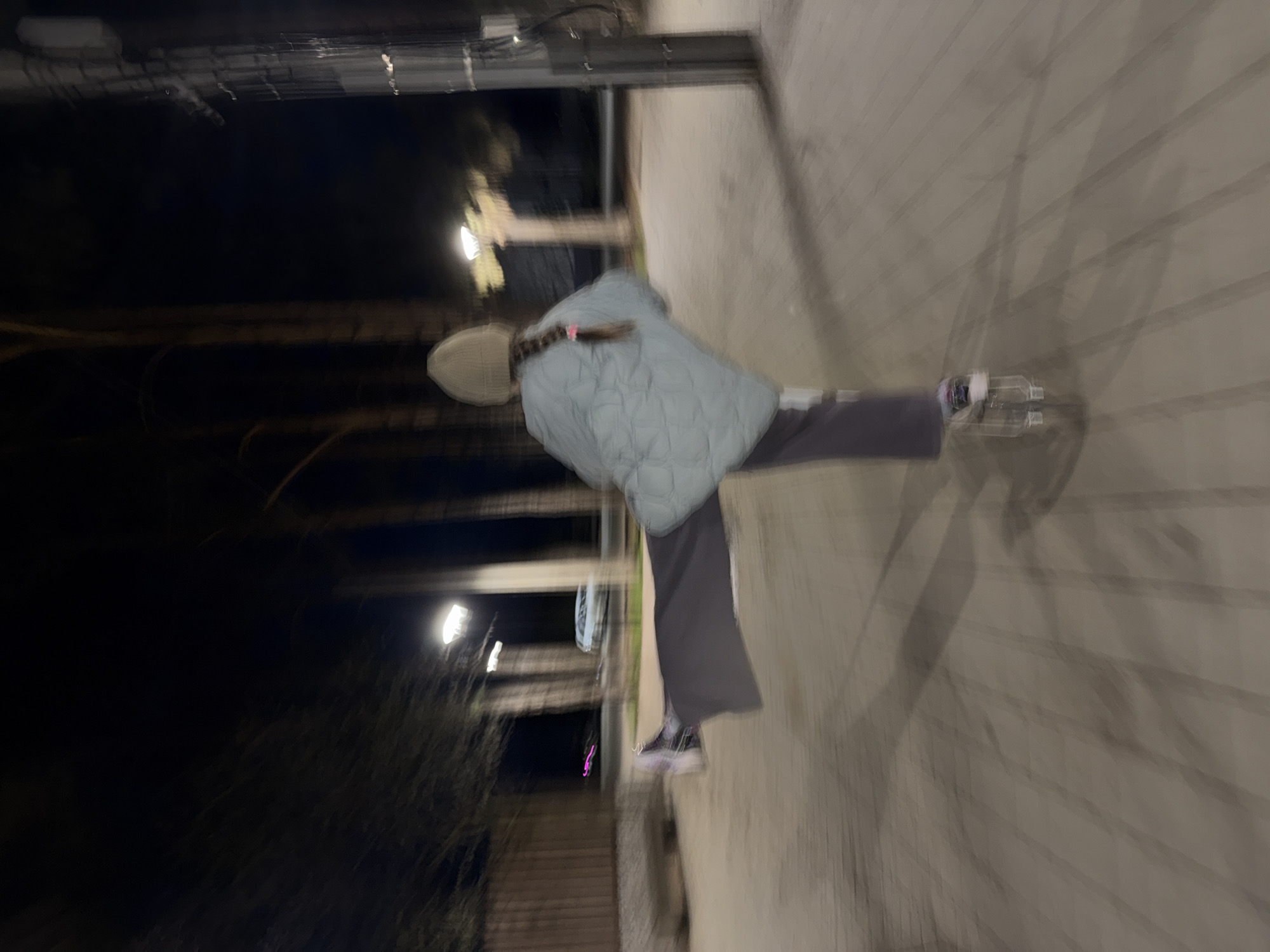 A person with a light-colored beanie, a braided hairstyle, and wearing a light blue quilted jacket and gray pants walks along a paved path at night. The path is lined with trees and streetlights are visible in the background. The image is slightly blurred, suggesting movement or a shaky camera. The location could be within the city of Votkinsk, Russia, given the prompt, but there is no concrete evidence in the image to confirm this.