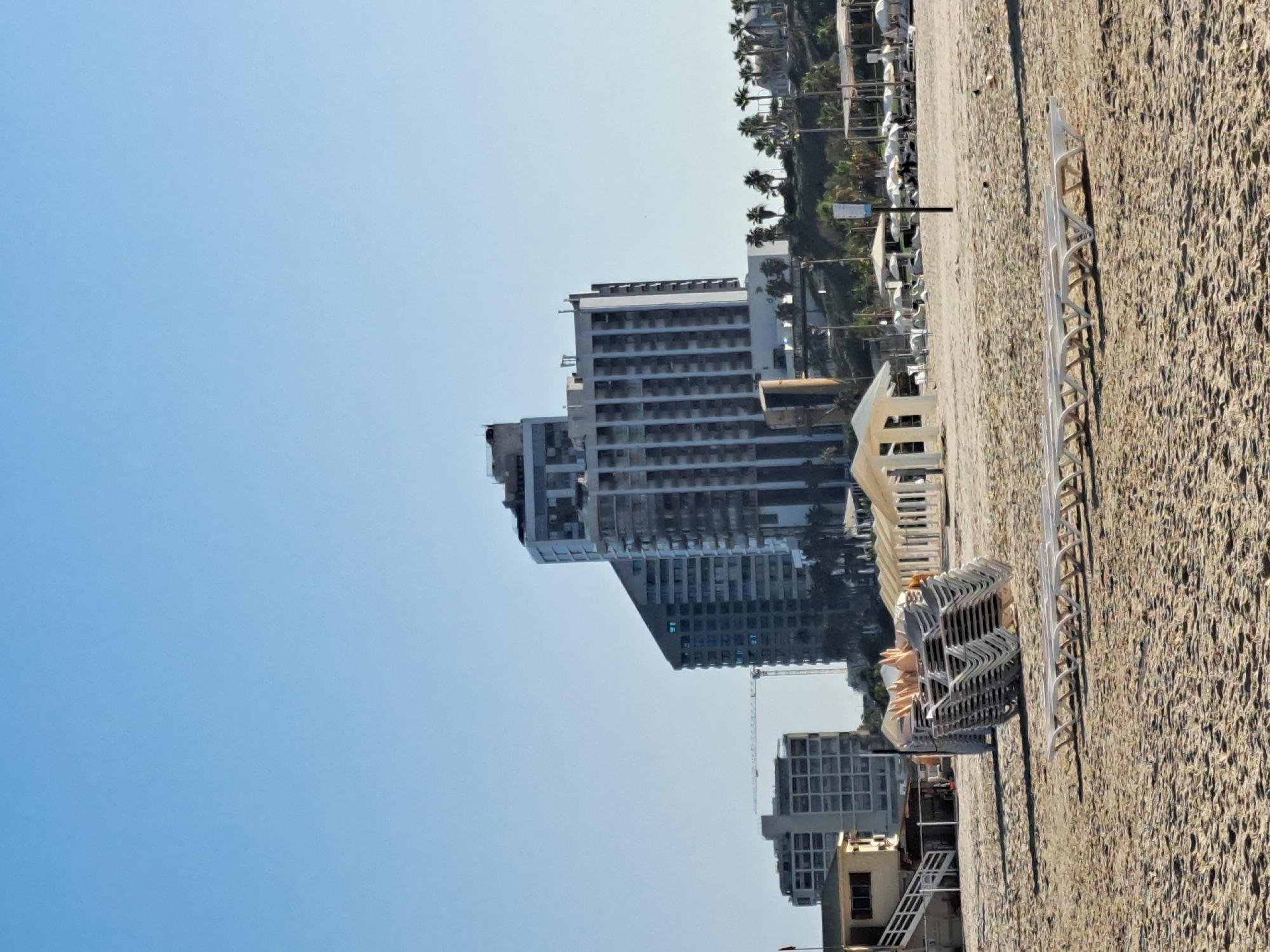 The image depicts a section of the beach in Herzliya, Israel. In the foreground, numerous white lounge chairs are arranged on the sandy beach. Some are stacked neatly, while others are spread out individually. In the middle ground, a large pile of stacked white lounge chairs is prominently visible, with a small lifeguard station or hut to the left. Further back, a row of white gazebos or shelters with umbrellas is set up. Beyond the beach structures, several modern high-rise buildings dominate the skyline, some featuring curved balconies and reflective windows. A construction crane is visible between two of the buildings, indicating ongoing development. Palm trees are interspersed throughout the scene, particularly around the buildings and along the coastline. The sky is a clear, pale blue, suggesting a sunny day. The overall environment is that of a developed coastal resort area.