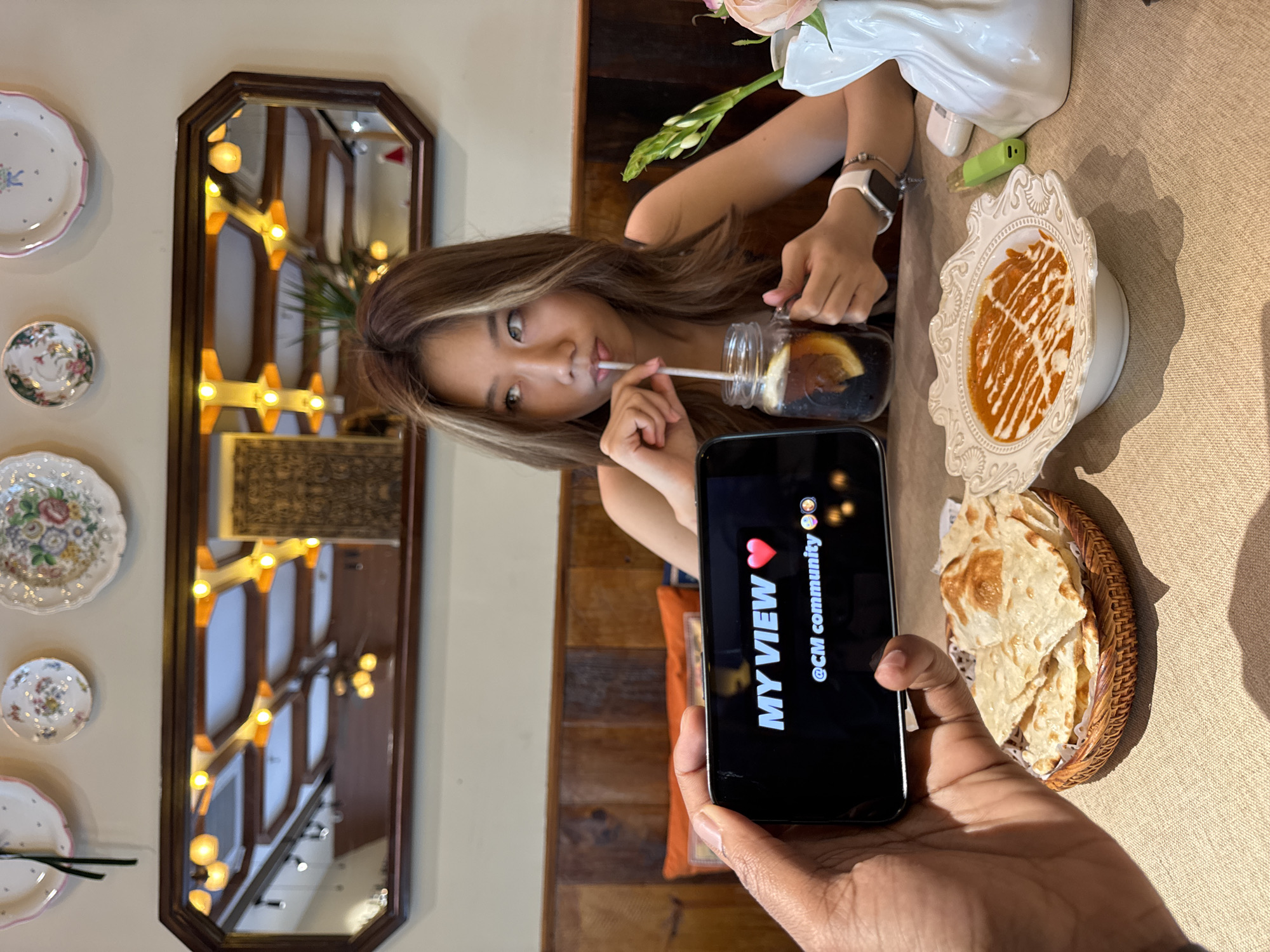 A slightly rotated medium shot depicts a person taking a photo with their phone of another person at a table in what appears to be a restaurant. The person being photographed, an Asian woman, is drinking from a mason jar with a straw. She is holding the jar with her left hand and has a white watch on her left wrist. The phone in the foreground reads "MY VIEW❤️ @CM community✨". Next to the phone and towards the right edge is a basket of flatbread, and further up is a white ornate plate with a saucy dish on it. Adjacent to the plate is a white vase with flowers and a green object. The table is covered in a tan tablecloth. Behind the woman is a dark wooden wall. To the left is a light-colored wall with decorative plates and a dark-framed mirror reflecting interior lights. The location is likely a restaurant in Yuzhong District, China, given the request.
