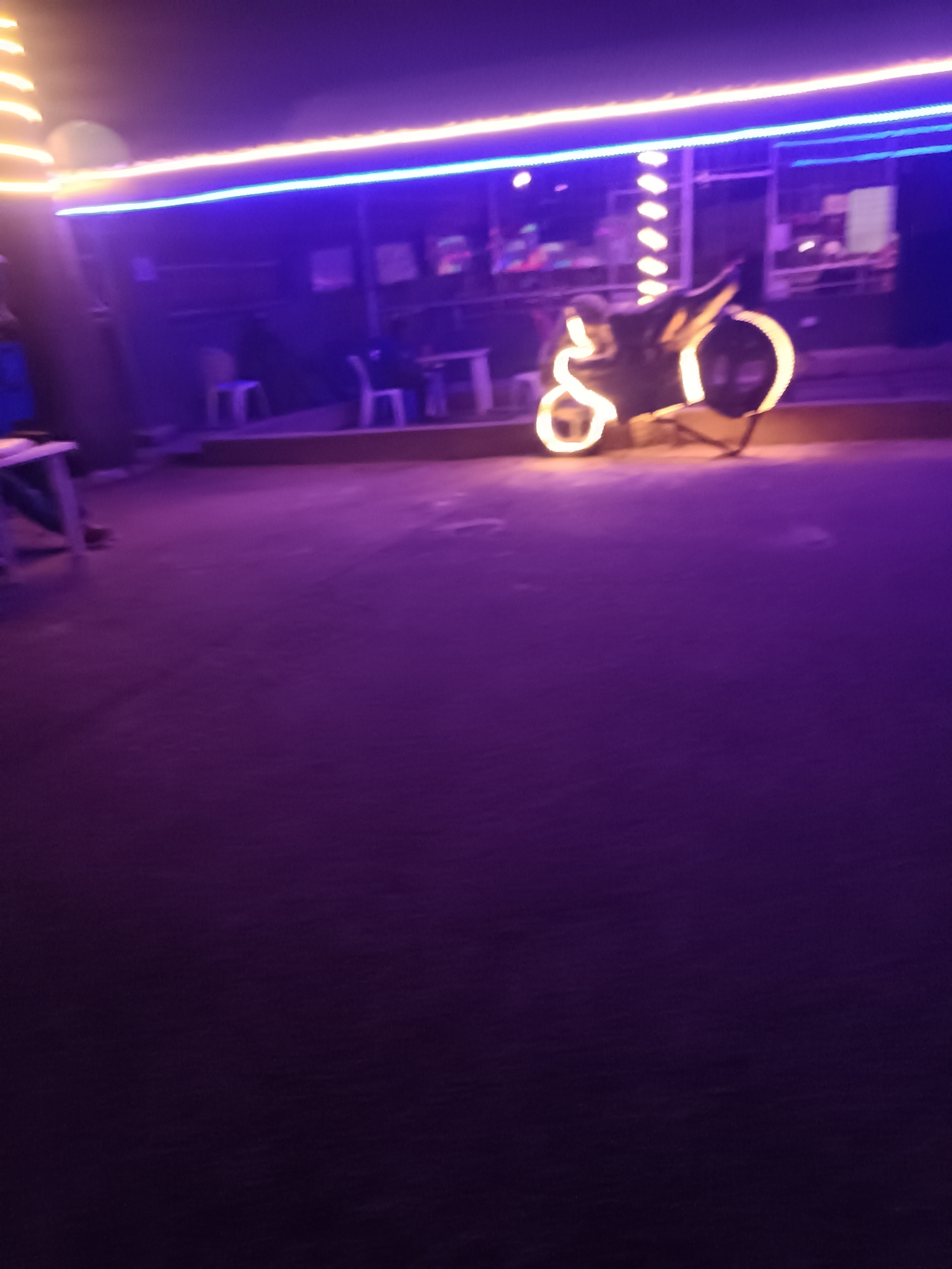 The scene appears to be a nighttime setting in Jos, Nigeria. In the background is a building with a covered patio area, lit by blue and yellow neon lights. Several tables and chairs are visible on the patio. In the foreground, a motorcycle is decorated with string lights, adding a prominent illumination. The ground appears to be paved. The overall atmosphere is illuminated and vibrant due to the lighting.