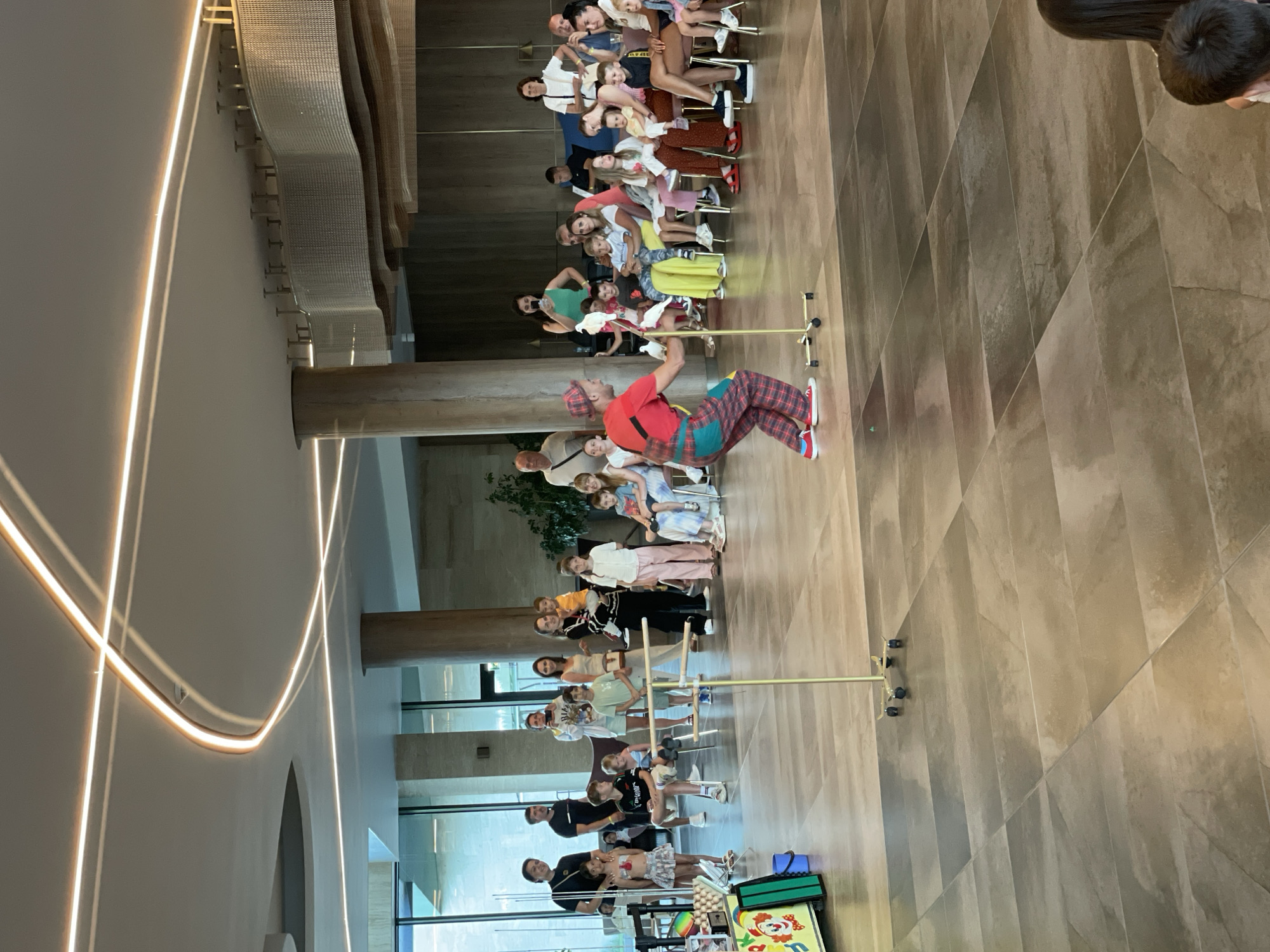 In Obzor, Bulgaria, a clown is performing for a crowd of onlookers, mostly children and adults, inside a modern building with high ceilings and contemporary lighting. The clown, dressed in red and white checkered overalls and a red shirt, is actively engaged in a magic trick, holding white doves. In front of the clown are two thin poles with horizontal bars, likely part of the performance. The audience is seated and standing, watching the show with interest. The background features large windows, architectural columns, and a textured ceiling with modern lighting fixtures. To the left of the performance area, a colorful sign depicting a clown is visible, along with a cart holding eggs, suggesting a children's entertainment event. The floor is tiled with large, rectangular, dark brown tiles arranged in a diagonal pattern.