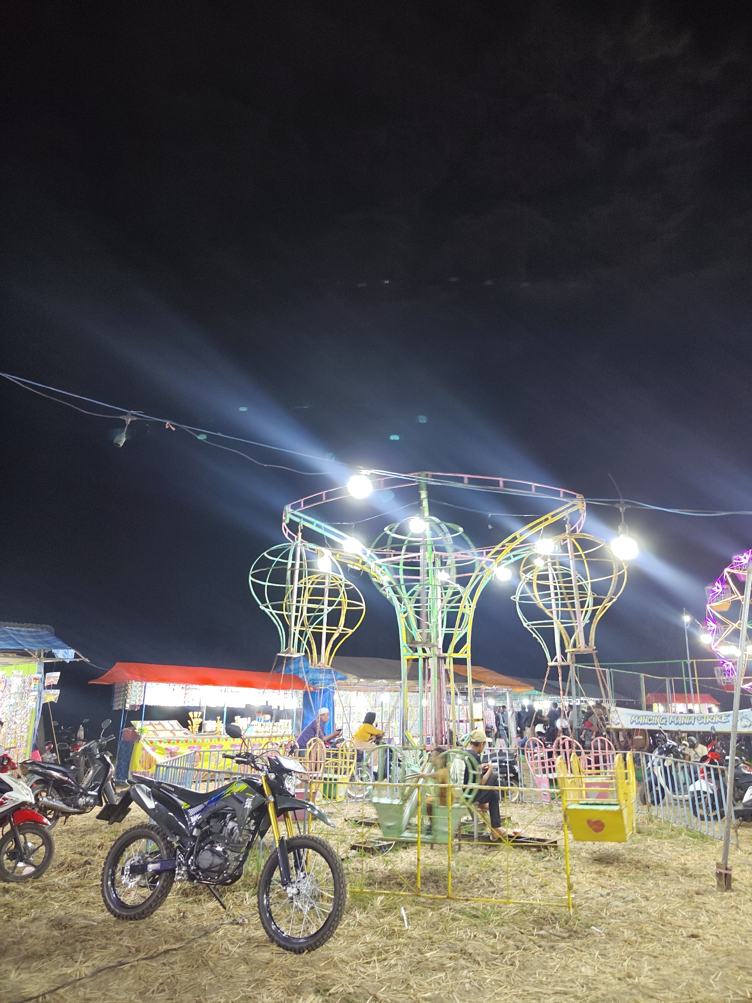 The image depicts a nighttime scene at what appears to be a local fair or amusement area in Suling Wetan, Indonesia. In the foreground, a black dirt bike is parked on dry, straw-like ground. Behind it, other motorcycles are visible, along with a red and white motorcycle to the left. The background features several brightly lit amusement rides. A Ferris wheel is visible on the far right, and a colorful, multi-tiered ride with balloon-like structures and hanging seats is prominent in the center. Stalls or vendors with red awnings are set up behind the rides. People are scattered throughout the area, some near the stalls and others appearing to be patrons of the rides. Overhead, strings of lights and wires crisscross the dark night sky, illuminated by the artificial light from the attractions. The overall atmosphere suggests a lively community gathering.