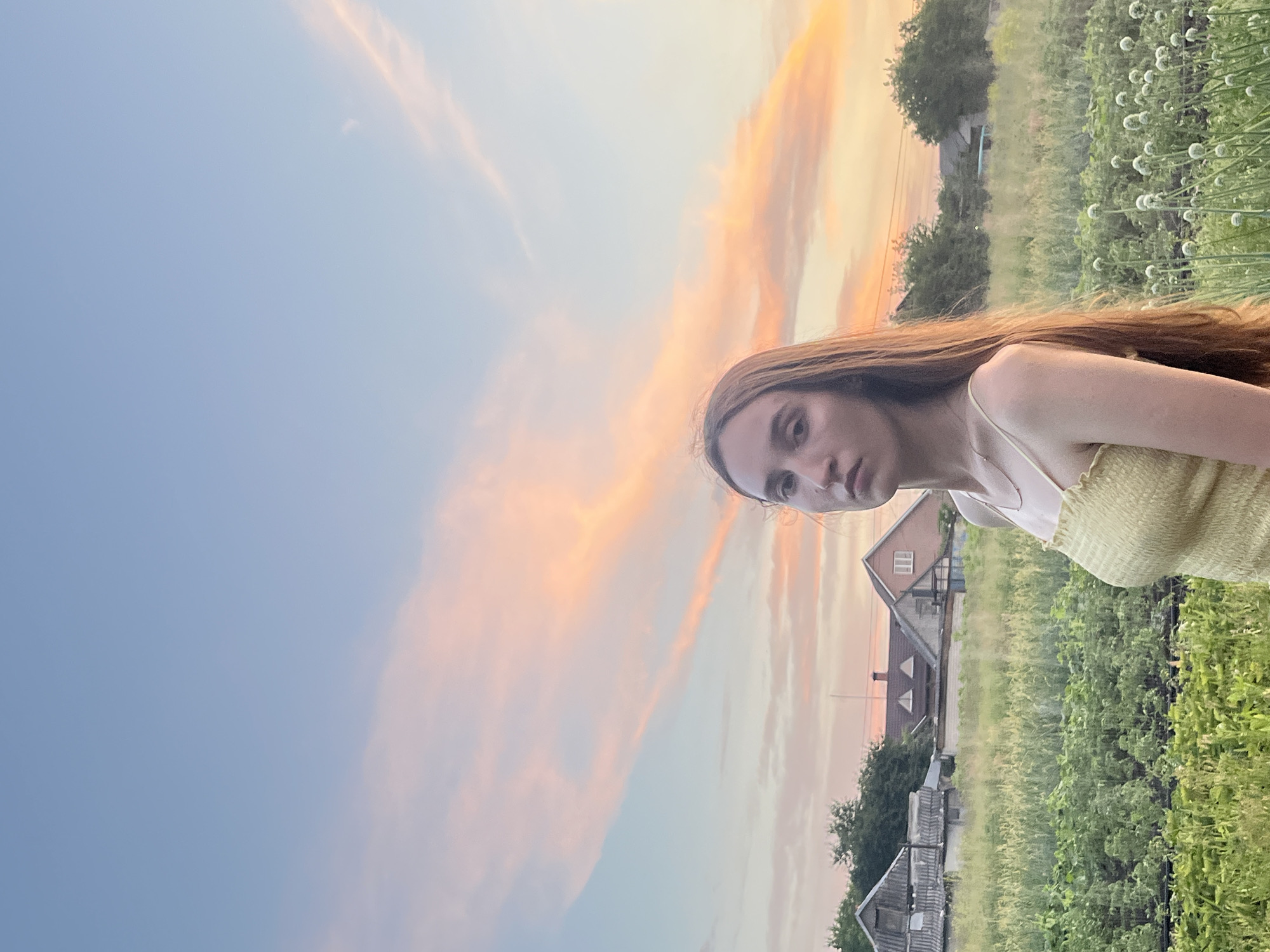 A young woman stands in a field with a village in the background. The sky is a soft blue with streaks of pink and orange clouds. The woman has long brown hair and is wearing a yellow dress. She is looking at the camera. The field is full of green plants, and there are some white flowers in the foreground. The village consists of a few houses and trees. The image appears to be taken at sunset.