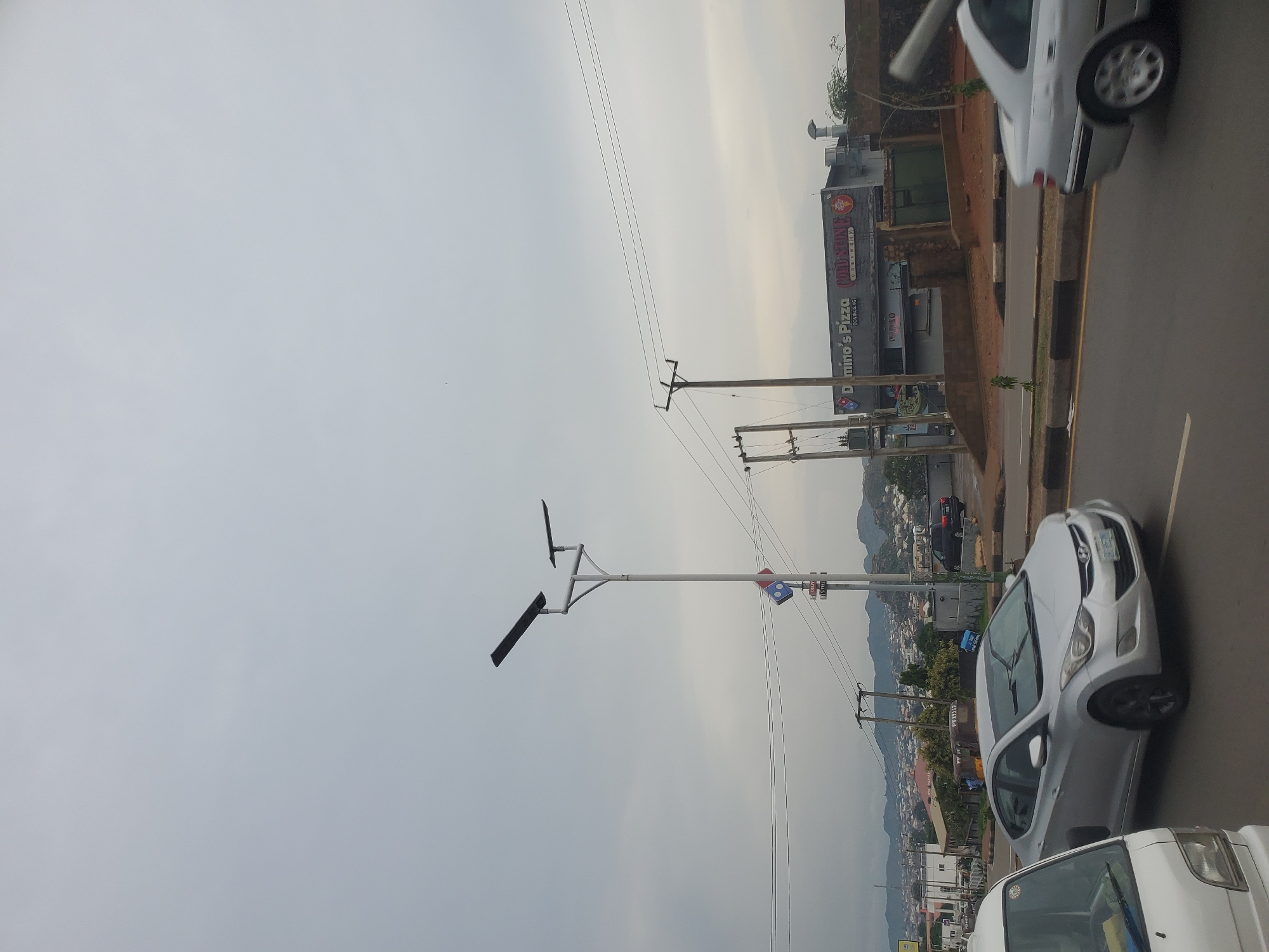 This image captures a street scene in Jos, Nigeria, under an overcast sky. In the foreground, a silver sedan is driving on a paved road, its front end slightly blurred due to motion. To its left, the front of a white vehicle, possibly a delivery van or small truck, is visible. In the mid-ground, a tall street lamp with two solar panels stands prominently. Attached to the lamp post is a Domino's Pizza logo. Behind the lamp post, a building houses a Domino's Pizza and a Cold Stone Creamery. Power lines stretch across the frame, supported by utility poles. In the background, a hilly landscape is dotted with residential buildings, suggesting an urban or semi-urban environment. The overall impression is a typical day in an African city with commercial establishments and residential areas coexisting.