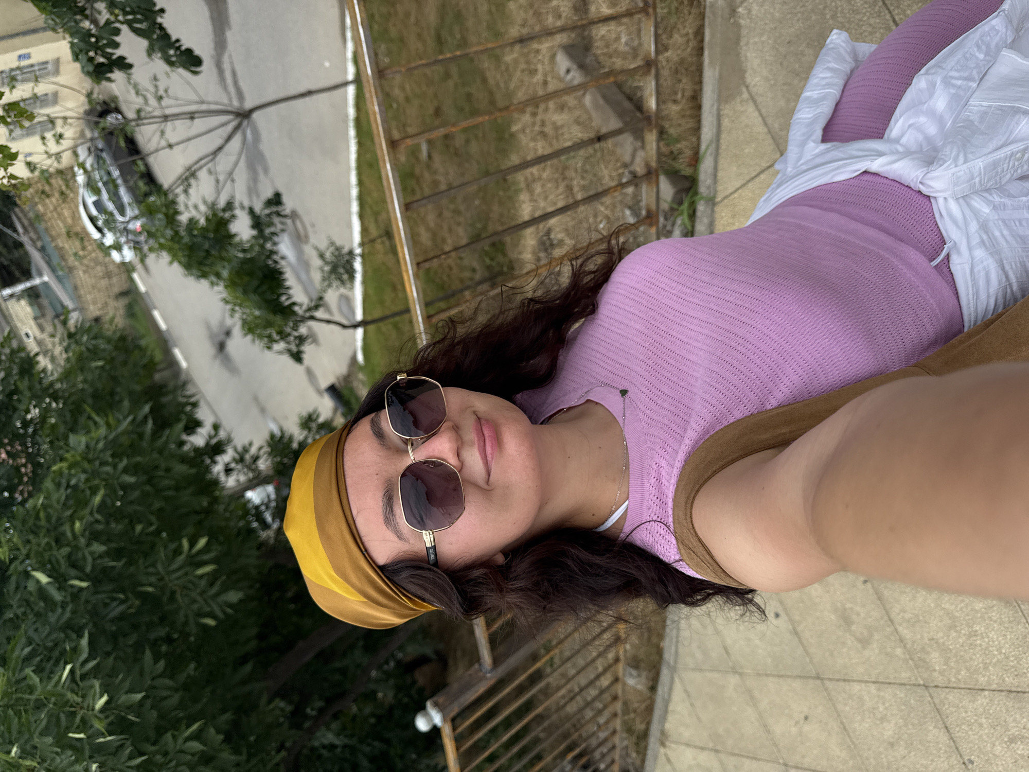 A young woman wearing a yellow and brown headscarf, large sunglasses, and a pink top is taking a selfie outdoors. She has long, dark, curly hair and is wearing a silver necklace with a small dark pendant. A white shirt is tied around her waist. In the background, there is a metal railing, trees, and a street with a parked car. The location is identified as Derbent, Russia. The overall scene suggests a casual outdoor setting.