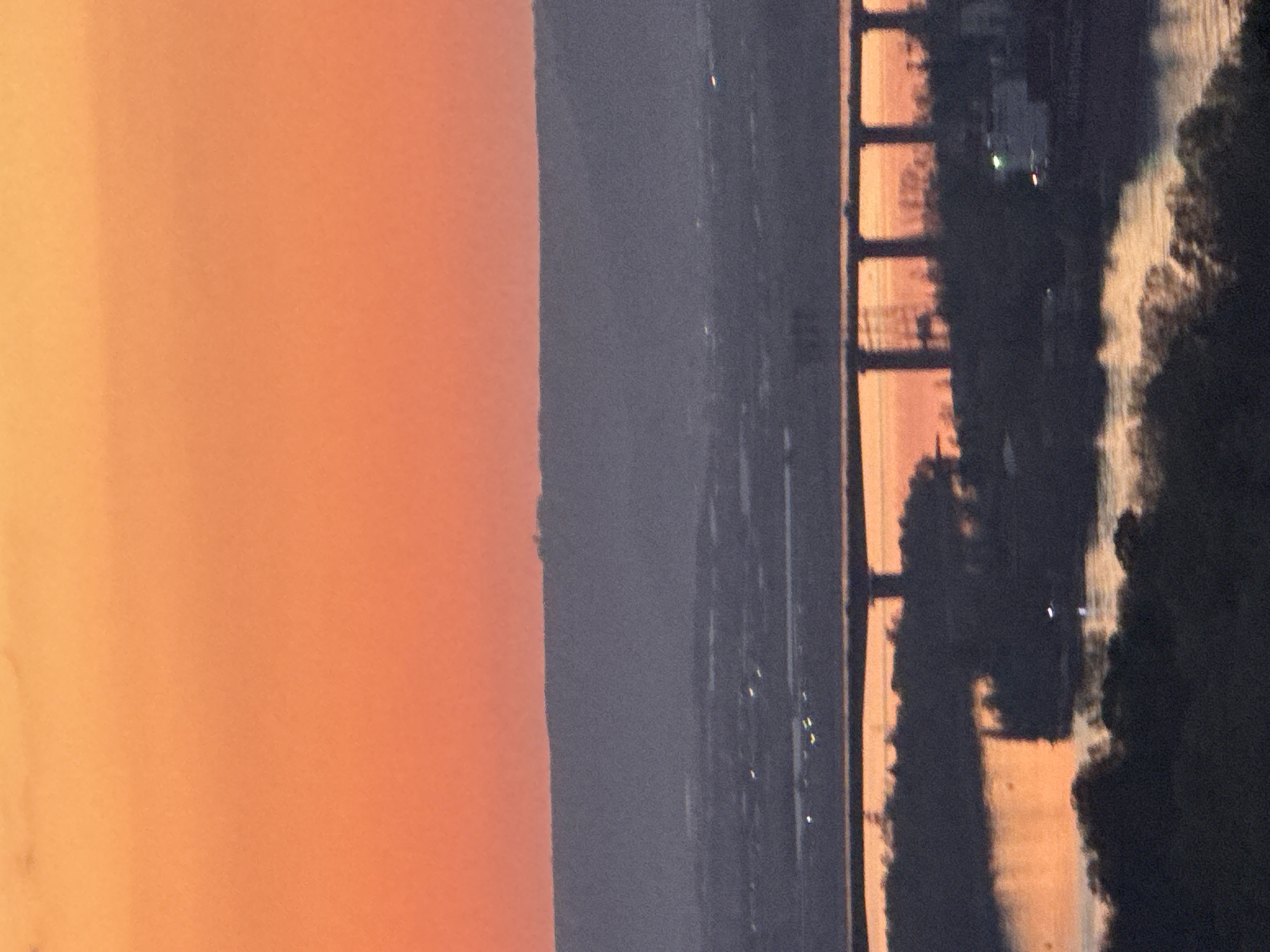 The image depicts a serene landscape scene captured during either sunrise or sunset, presenting a dramatic interplay of light and shadow over a body of water and distant land. The sky, occupying the left portion of the frame, displays a warm, soft gradient of colors, transitioning from lighter yellow-orange to deeper orange and reddish-pink hues, characteristic of golden hour lighting.
Horizontally across the middle of the image, a dark, silhouetted stretch of distant land or hills separates the vibrant sky from the foreground elements. Faint, scattered points of light are visible within this dark landmass, suggesting distant urban structures or activity.
On the right side, a prominent bridge structure is silhouetted against the warm light. Its design features a series of regularly spaced vertical and horizontal elements, indicative of a large infrastructure project spanning a significant body of water. The water beneath the bridge and in the lower right quarter of the image is textured and appears to be rippling, beautifully reflecting streaks of the warm orange and yellow light from the sky amidst darker areas. Dark, irregular shapes, likely trees or dense vegetation, are also silhouetted along the water's edge and near the bridge.
The scene is tranquil and devoid of specific human activity, focusing instead on the natural beauty and the architectural presence of the bridge. The time of day is clearly either dawn or dusk due to the distinct warm lighting. There is no visible text. Given the location context of Varna, Bulgaria, the bridge is likely the Asparuhov Bridge over Lake Varna.