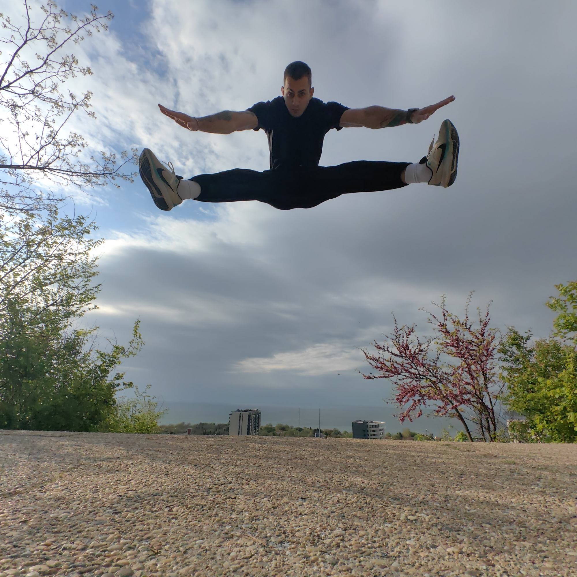A male athlete is captured mid-air, performing a dynamic jump split outdoors. He wears a black t-shirt, black pants, white socks, and light-colored athletic shoes with a dark logo. His arms are stretched wide horizontally, mirroring his legs which are extended in a full split, showcasing remarkable flexibility and strength. His gaze is directed intently towards the viewer.
The scene unfolds under a partially cloudy sky, revealing patches of blue amidst the white and gray clouds, indicating daytime. On the left, branches of a tree with fresh green leaves extend into the frame, while on the right, a tree with distinctive pink blossoms adds a touch of color.
In the mid-ground, below the foliage, several modern buildings are visible on a distant ridge, overlooking a vast body of water, which is the Black Sea, consistent with the location context of Varna, Bulgaria. The foreground consists of a textured ground covered in small, light-colored pebbles. The low camera angle emphasizes the height and vigor of the man's jump against the expansive sky and distant landscape. There is no visible text in the image.
