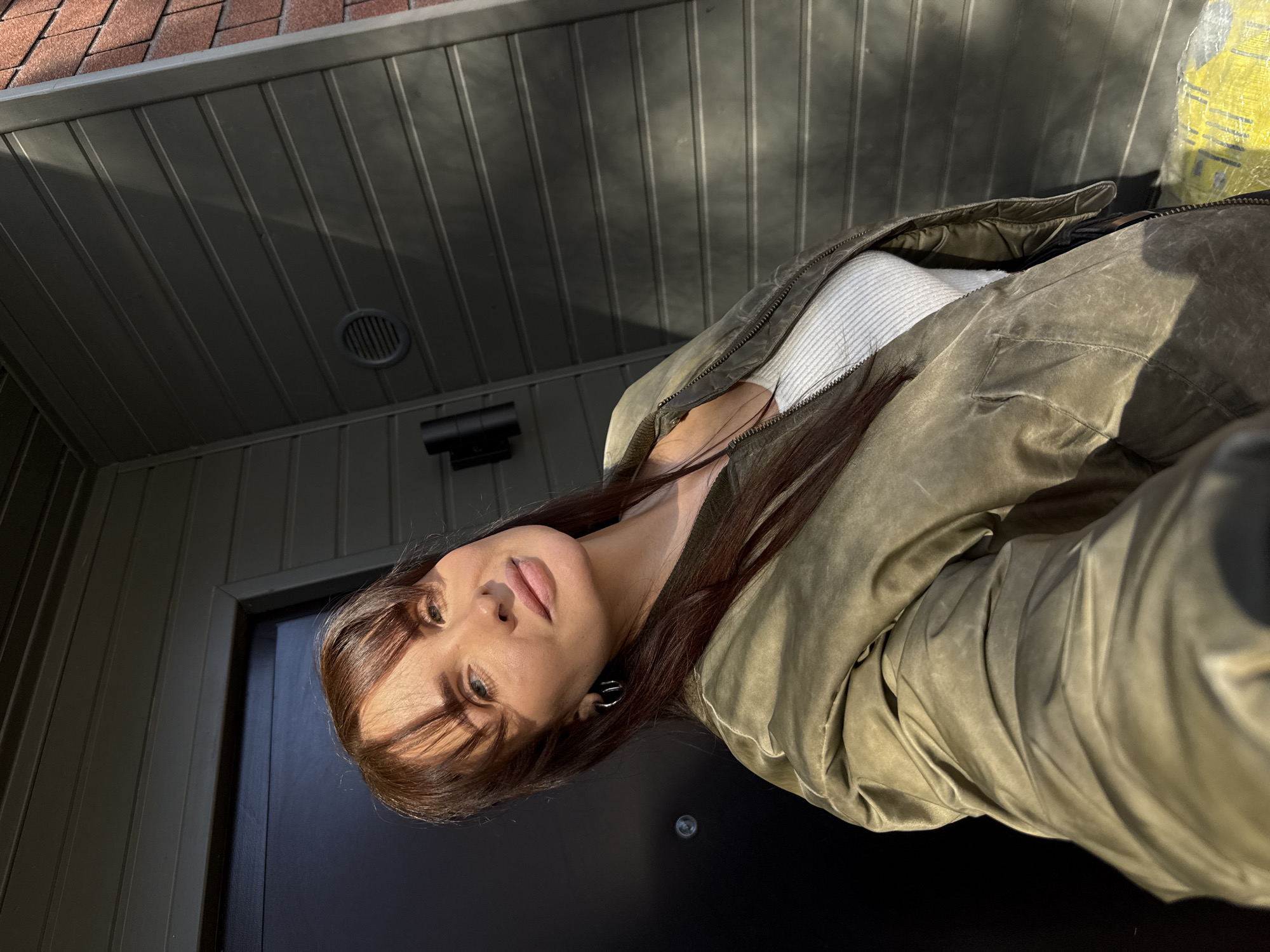 A woman with long brown hair and bangs, wearing a white top and an olive green jacket, is standing in front of a black door. The door is set within a covered entryway with a gray ceiling and dark gray walls. The roof, visible in the upper left corner, is made of red tiles. The photo appears to be taken in Chaikovsky, Russia.