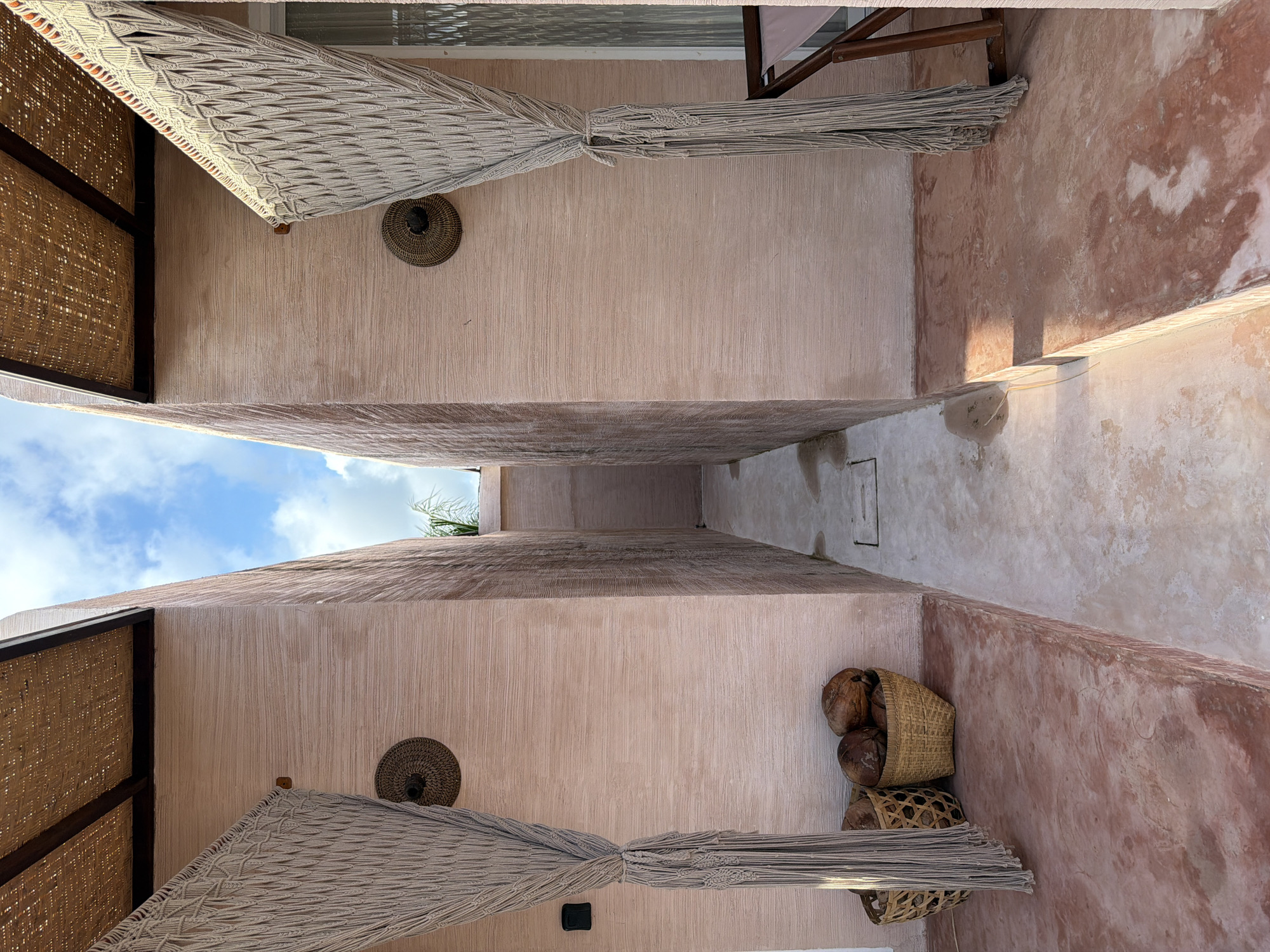 Captured from a low angle, the photograph presents a narrow outdoor passage between two buildings in North Kuta, Indonesia. The facades of the structures exhibit a textured, light pink finish, contrasting with patches of red paint. Woven awnings protrude from the buildings' sides, providing shade. Decorative straw baskets adorn the walls, and baskets filled with coconuts are arranged on one side. The sky, visible through the passage, features scattered clouds.