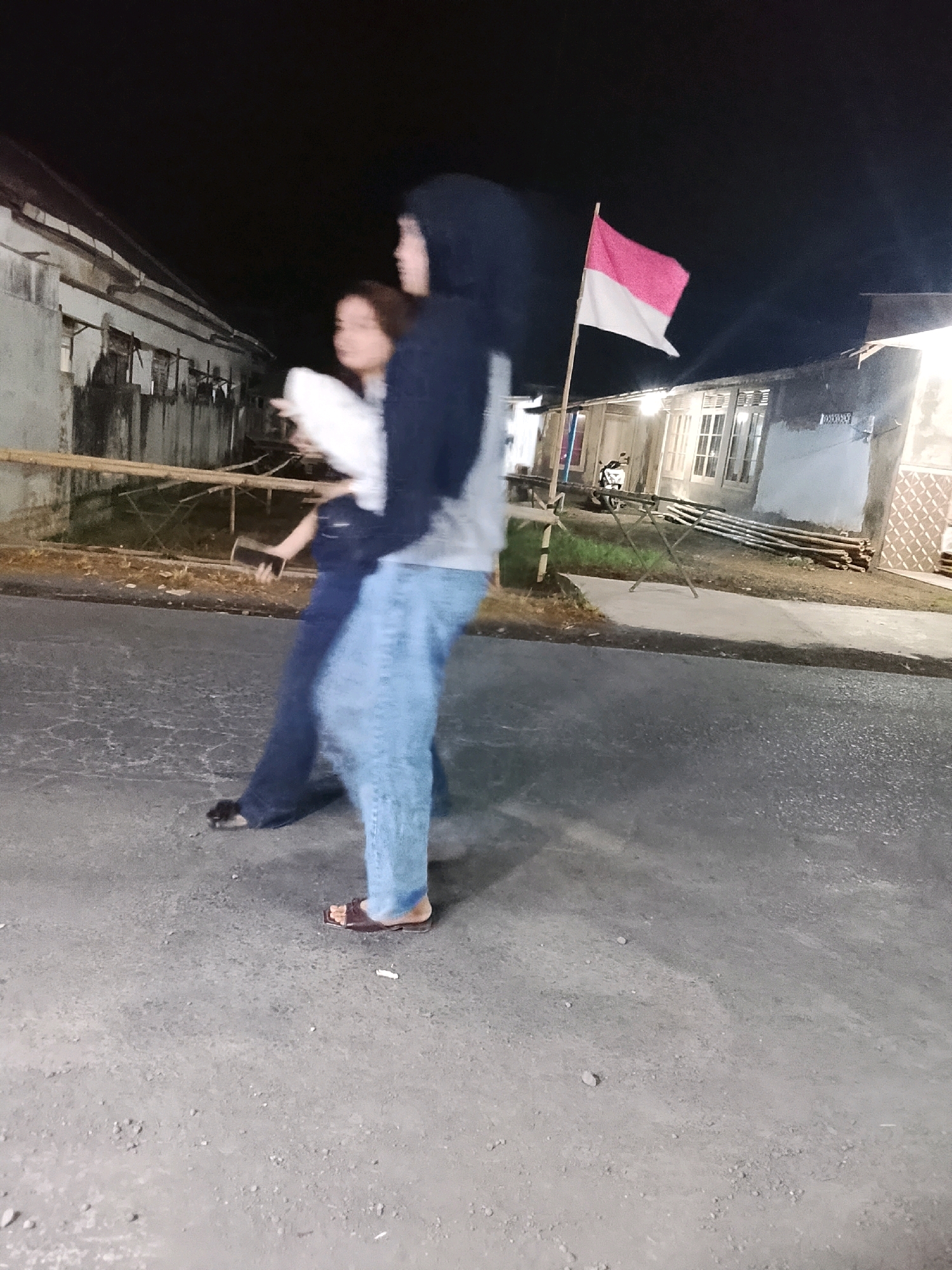 In the city of Sukorejo, Indonesia, at night, two individuals are captured in a photograph. The person in the foreground, wearing a black hooded sweatshirt and light blue jeans, is positioned in the center, facing away from the camera and slightly to the right. This individual is holding another person, who is facing the camera and partially obscured. The person being held wears a white top and dark pants. Both individuals are walking on a paved road. To the left of the frame, a building with a light-colored wall and a sloped roof is visible. A bamboo structure runs horizontally in front of the building. A flag, with red on top and white on the bottom, is prominently displayed on a pole to the right of the individuals. Further to the right, another building with windows and a patterned wall is observed. A motorcycle is parked near the building on the left. The overall scene is dimly lit, suggesting it is nighttime.