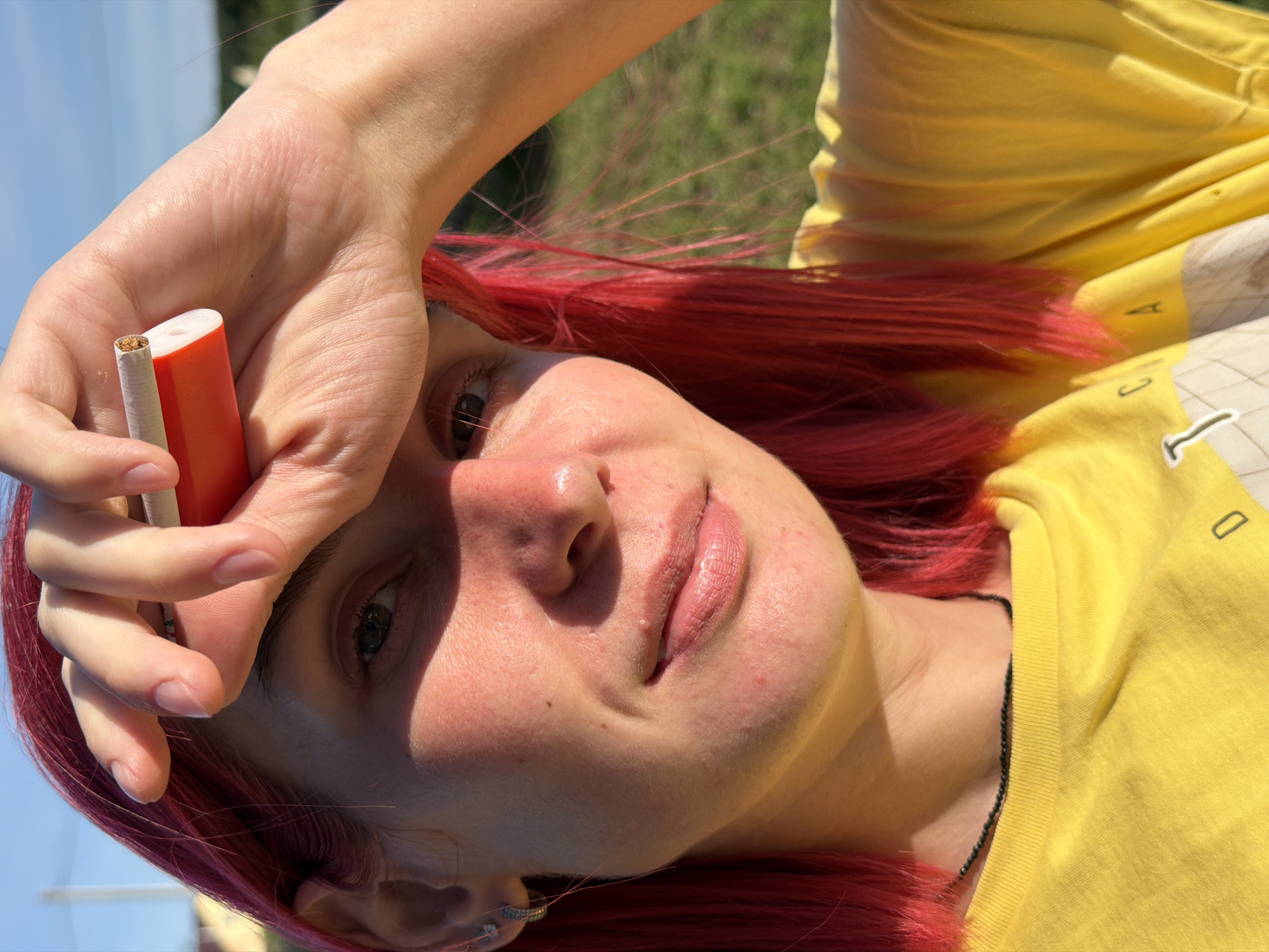 This media file shows a close-up, head-and-shoulders shot of a person with bright red hair. The person is wearing a yellow t-shirt and has their right hand raised to their forehead, holding a cigarette and what appears to be an orange vape pen. The person's face is tilted upwards, and their eyes are open, looking towards the camera. Sunlight is casting a shadow on the person's forehead and cheek.
The background is blurred, but appears to be an outdoor setting with green grass and a blue sky. The lighting suggests it is daytime, likely a sunny day.
Notable details include the contrast between the person's red hair and the yellow shirt, the detail of the cigarette and vape pen, and the texture of the person's skin. The emotion conveyed is somewhat ambiguous due to the angle and the objects being held, but the open eyes and direct gaze suggest a moment of awareness or perhaps defiance. No visible text is present in the image. The location context is Chaikovsky, Russia.