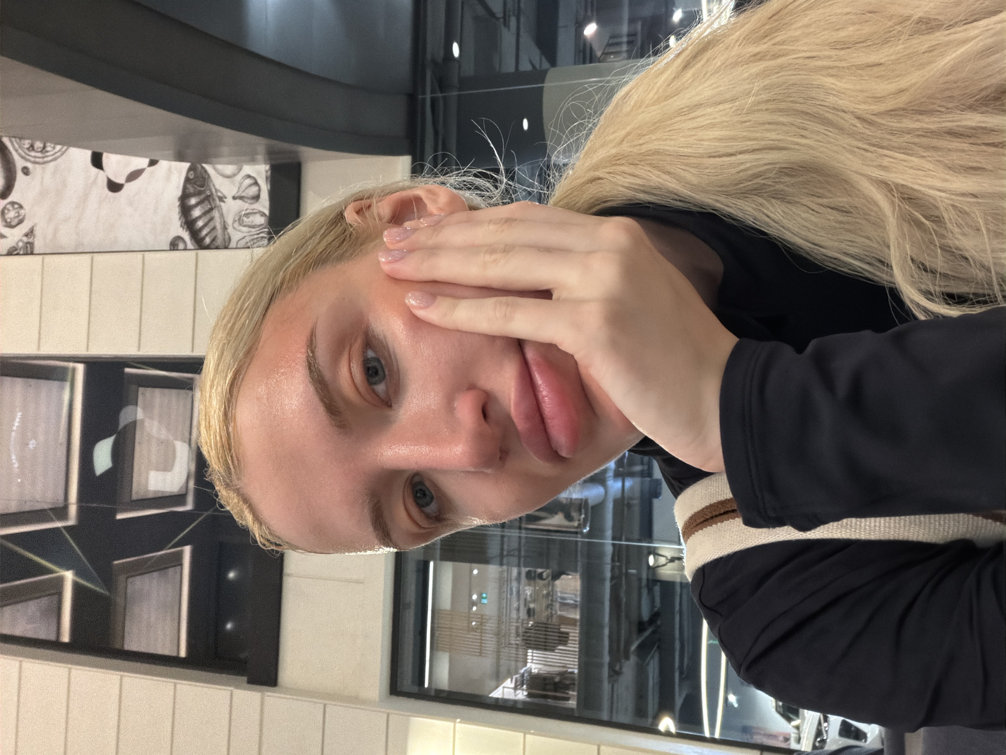 A fair-skinned woman with blonde hair pulled back in a ponytail rests her face in her hand. She has light blue eyes and prominent lips. She wears a dark top with a striped strap. The background is a commercial interior, possibly a restaurant or cafe, with tiled walls and framed artwork. Through a large window, an interior scene with bright lighting and equipment suggestive of a commercial kitchen or food preparation area is visible. The image lacks information to ascertain the exact location in Belgrade, Serbia.