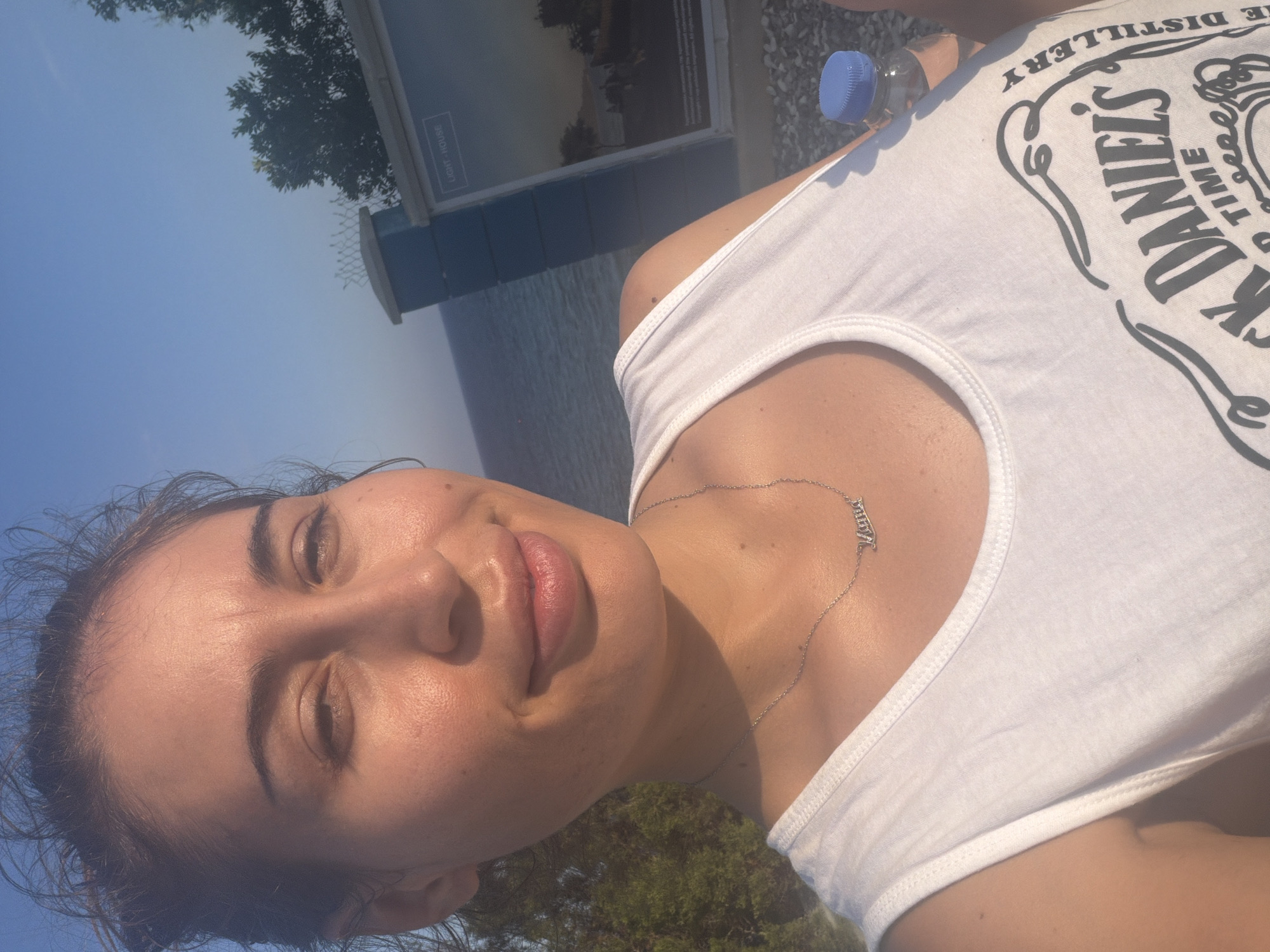A selfie is taken outdoors in Pervolia, Cyprus. The subject, a young woman with dark hair pulled back, is wearing a white tank top with a "Jack Daniel's" logo. She has a silver necklace with the name "Mama" on it. Behind her, the sea is visible, along with some trees and a building with a sign that reads "LIGHT HOUSE." The sun is shining, casting a warm glow on her face.