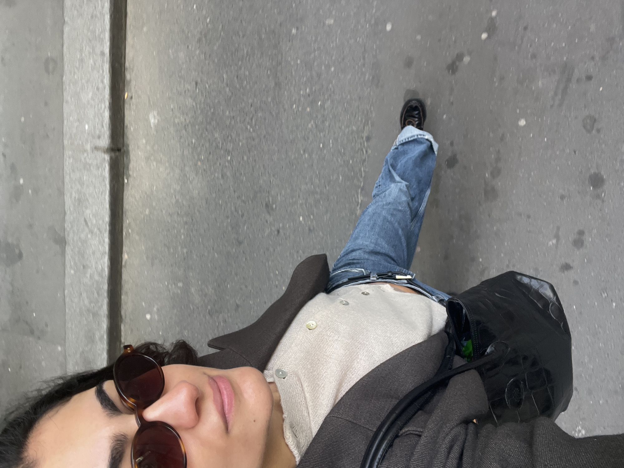 A high-angle selfie captures a person from an overhead perspective, looking upwards towards the camera, suggesting they are holding the camera themselves. The individual has dark, wavy hair and wears tortoiseshell-patterned sunglasses positioned on their nose. Their neutral expression reveals the lower part of their face, including their lips.
The person is dressed in a light-colored, possibly beige or off-white, knitted top with several buttons, layered under a dark brown or charcoal-grey coat. They are wearing blue jeans, cuffed at the bottom, with a black belt, and dark shoes. One leg is extended forward, implying motion or a pose while walking or standing on a paved surface, likely a street or sidewalk in an urban setting like Paris, France. A black handbag, possibly with a textured or crocodile-embossed finish, rests on the coat. The ground is grey, consistent with asphalt or concrete, featuring a distinct vertical line on the left, possibly a curb or building edge. The lighting appears to be natural daylight, possibly overcast. No visible text is present in the image.