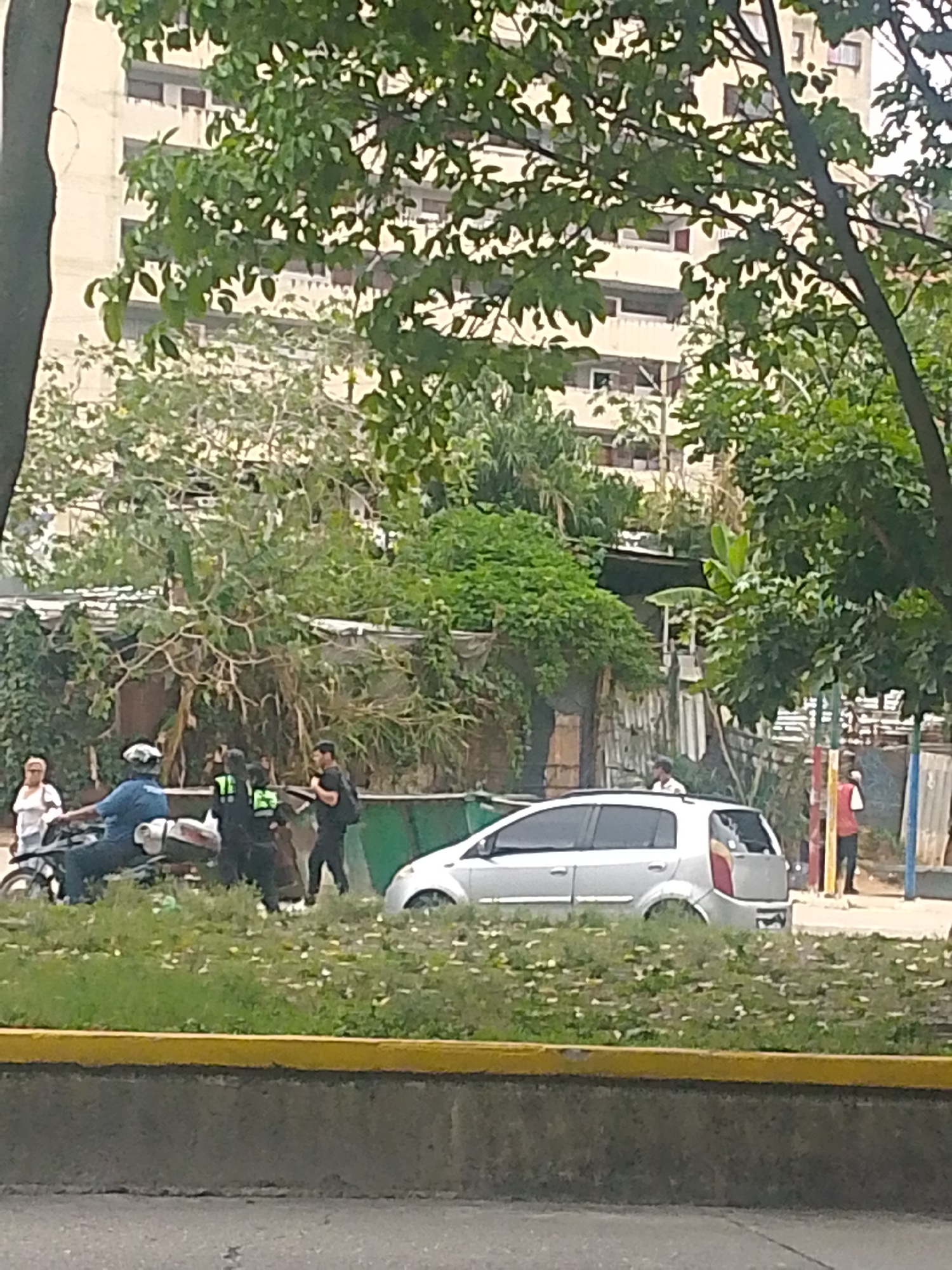 The image shows a scene in what appears to be Caracas, Venezuela. In the foreground is a low concrete wall with a yellow strip running along the top. Behind the wall is a patch of green grass and weeds.
A silver compact car is parked on the side of the road. A motorcycle is positioned beside the car, occupied by a person wearing a blue shirt and a white helmet. Two people stand near the motorcycle; one is wearing what appears to be a green vest with a number on it, and the other is dressed in black, carrying a backpack. Another person, dressed in a white shirt, stands on the left of the motorcycle.
In the background, behind a fence and some vegetation, a shanty can be seen. Further back, there is a multi-story building visible through the trees. A playground area is also visible on the right side of the image, where two more individuals are standing.
Overall, the scene appears to be a street with a mix of urban and informal elements, with a variety of people and objects.