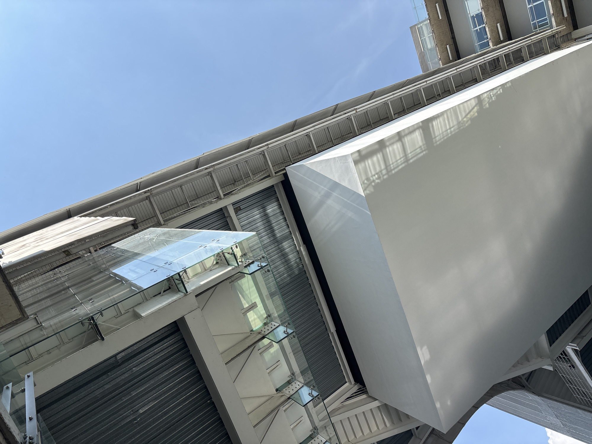 The image presents an upward view of modern architectural structures, likely located in Mexico City, Mexico. The sky, a clear blue, provides the backdrop for the buildings.
On the right, a large, white, cuboid structure dominates a portion of the frame. Reflections and shadows play across its surface. To the left, a glass-enclosed walkway or bridge is visible, supported by white frames and affixed with visible metal fasteners. The reflective glass offers glimpses of the sky above. Further to the left, corrugated metal is visible. A partial view of another building with windows can be seen toward the upper center.