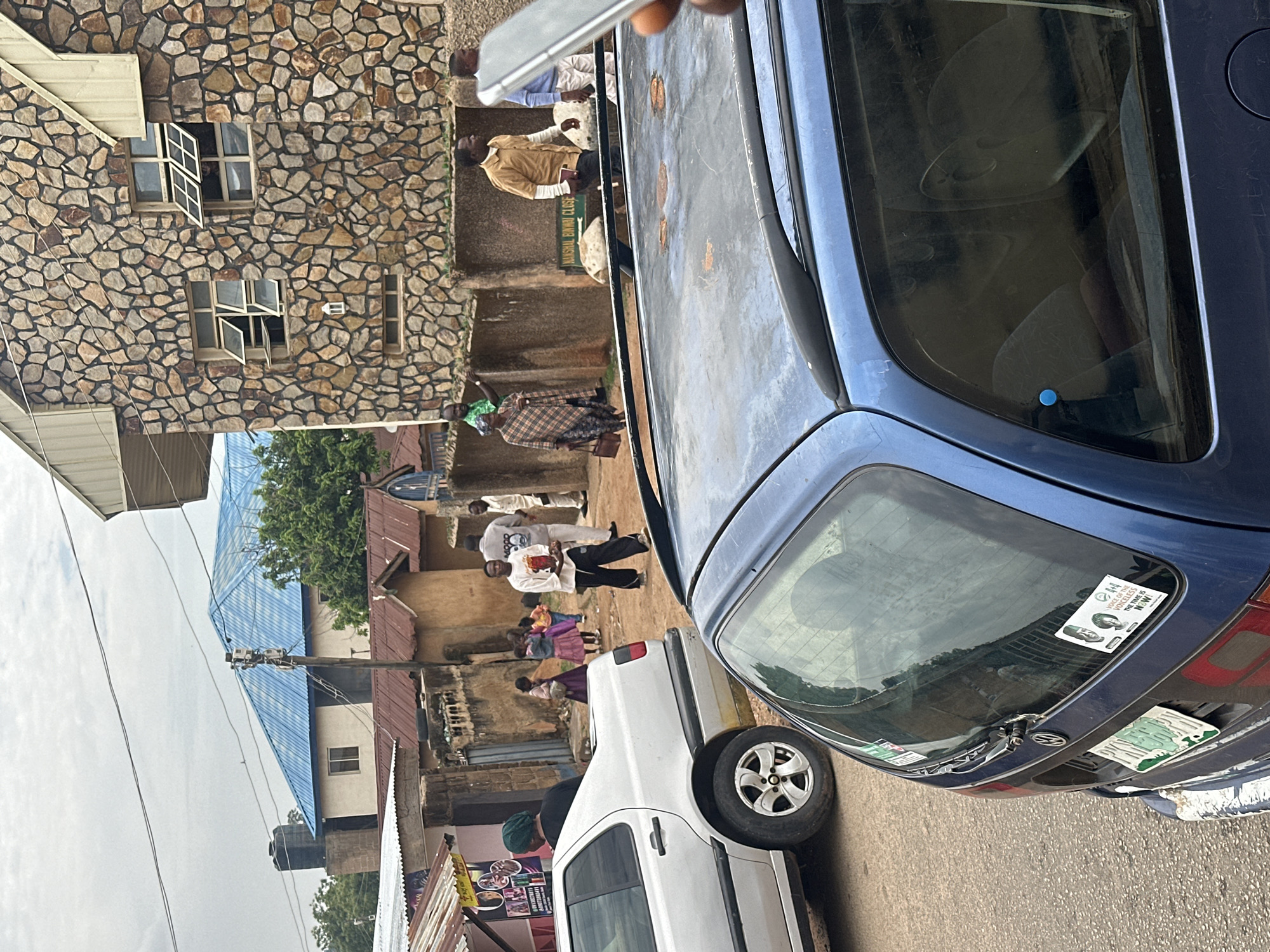 This image depicts a bustling street scene in an urban residential area of Unknown, Unknown, under an overcast sky.
In the immediate foreground, a blue Volkswagen station wagon is partially visible, mainly its rear and the front portion of its roof, which shows several patches of rust. A hand holding a silver smartphone is prominent in the extreme right foreground, partially obscuring the car's roof and the middle ground. The rear window of the blue car features a white sticker with two depicted individuals and the text "VOICE OF THE VOICELESS THE TIME IS NOW!". A green license plate is visible on its rear.
To the left, a silver Volkswagen sedan (resembling a Jetta or Vento) is parked on the dirt road, facing away from the viewer. A woman wearing a green head covering and a dark top stands near the rear of this car.
The middle ground is populated by several individuals. A group of children, predominantly young girls in colorful dresses (one in purple), are visible further down the street near some smaller buildings. Walking from left to right, a man in a tan long-sleeved shirt carries a dark book or notebook. To his right, a man in a white long-sleeved shirt with the word "MOOD" printed on it walks away from the viewer. Further left, a woman in a brown and white patterned wrap dress walks towards the left, with a man in a green shirt partially visible behind a low concrete wall.
On the right side of the frame, a prominent two-story building is constructed with a distinctive facade of irregularly shaped, multi-toned natural stone. It features multiple rectangular windows with white frames; some upper windows are open outwards. A green and white street sign bearing the inscription "NANSHAL BINWAI CLOSE" is affixed to a low concrete wall in front of this stone building.
In the background, overhead utility wires crisscross the sky. A building with a bright blue corrugated metal roof stands out, partially obscured by another building to its left that has a black water tank elevated on a concrete structure on its roof. Closer to the middle ground, several single-story structures with reddish-brown corrugated metal roofs line the dirt road, suggesting a mix of residential and small-scale commercial activity. Green foliage is visible in several areas, including behind buildings and near a utility pole.